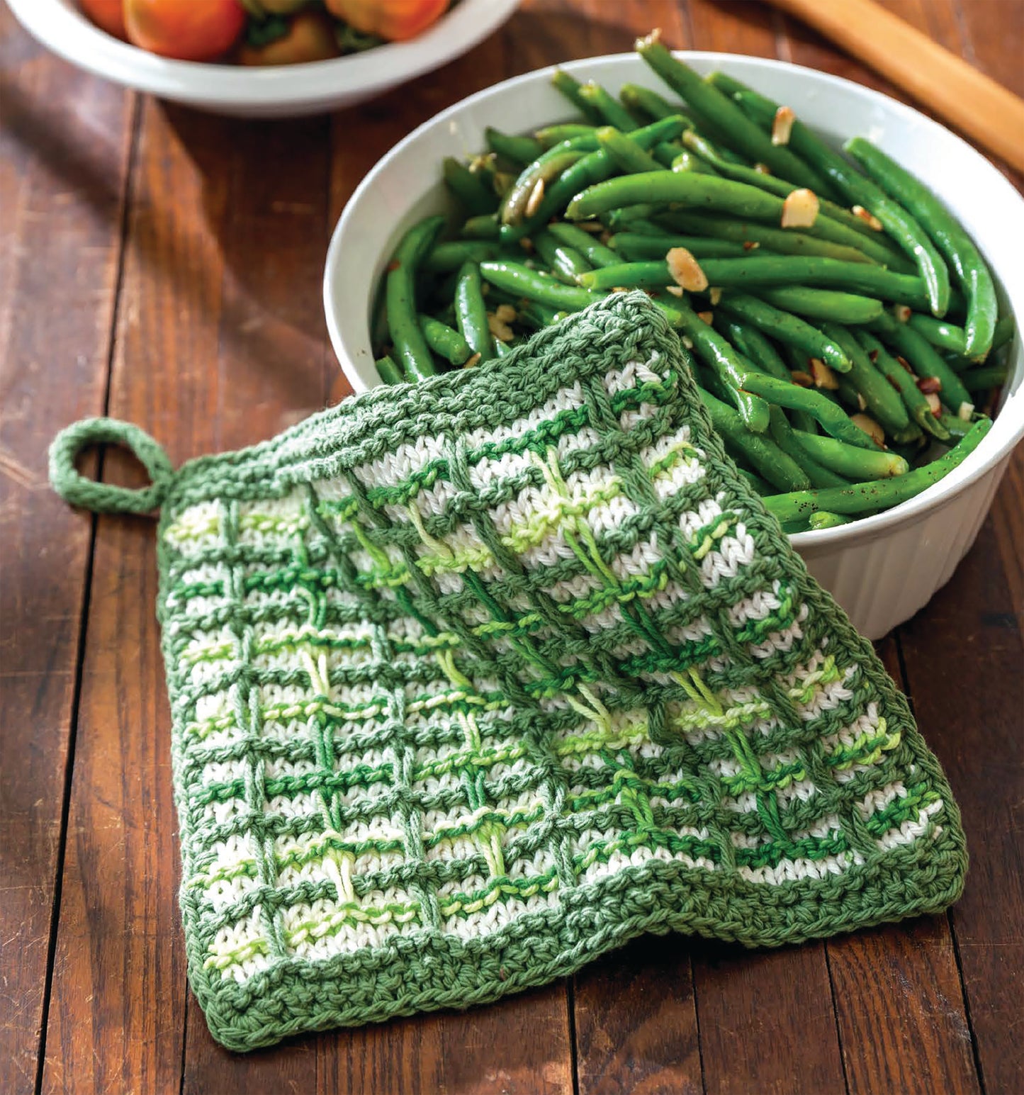The Make in a Weekend Potholders and Dishcloths by Leisure Arts, shown here in green and white on a wooden table beside a bowl of green beans and garlic, is perfect for adding handmade charm to your kitchen. Tomatoes can be seen in the background.