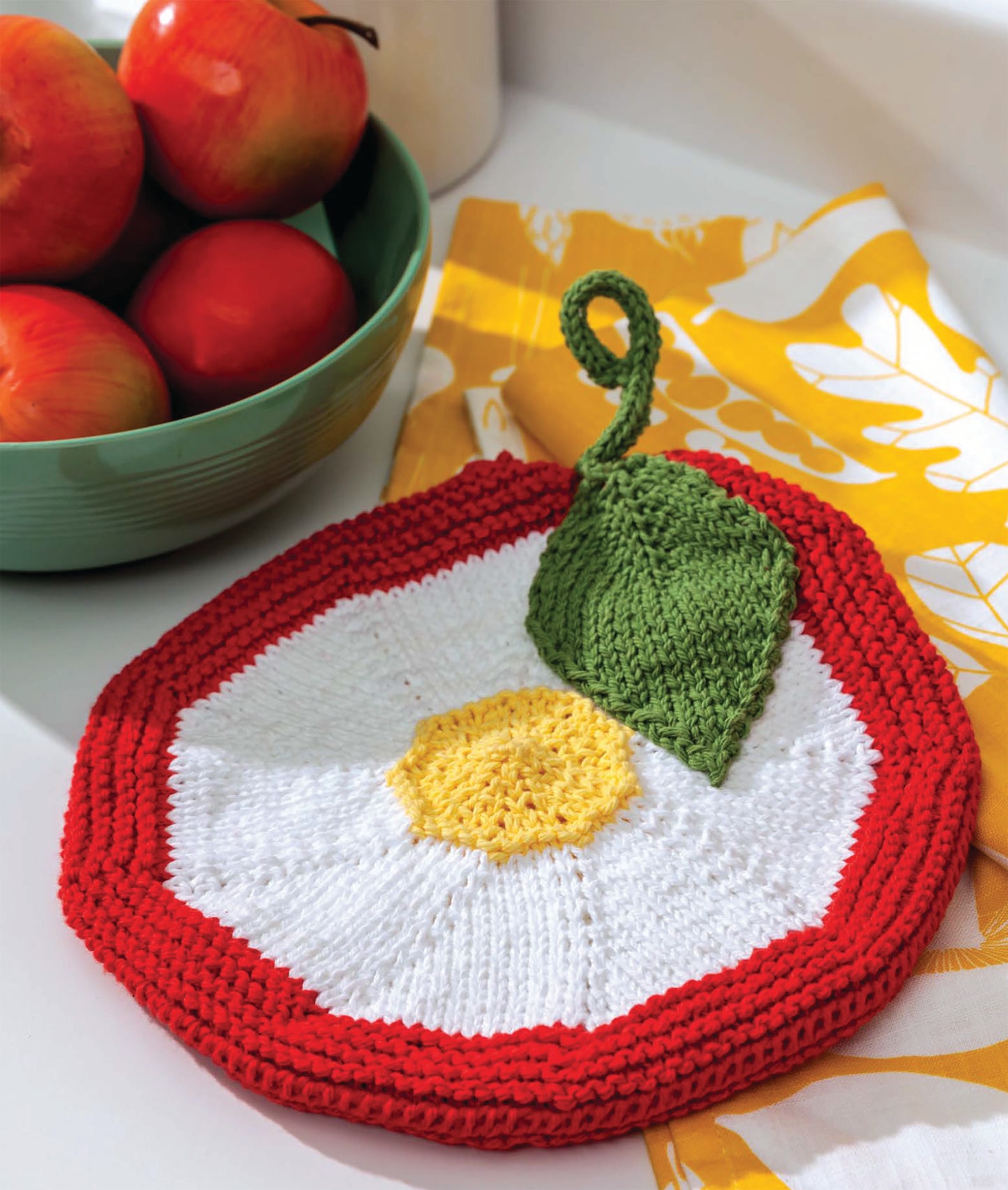 The Make in a Weekend Potholders and Dishcloths by Leisure Arts features a round, apple-themed knitted potholder with red border, white center, yellow core, and green leaf—adorable kitchen decor displayed on a yellow patterned cloth near apples.
