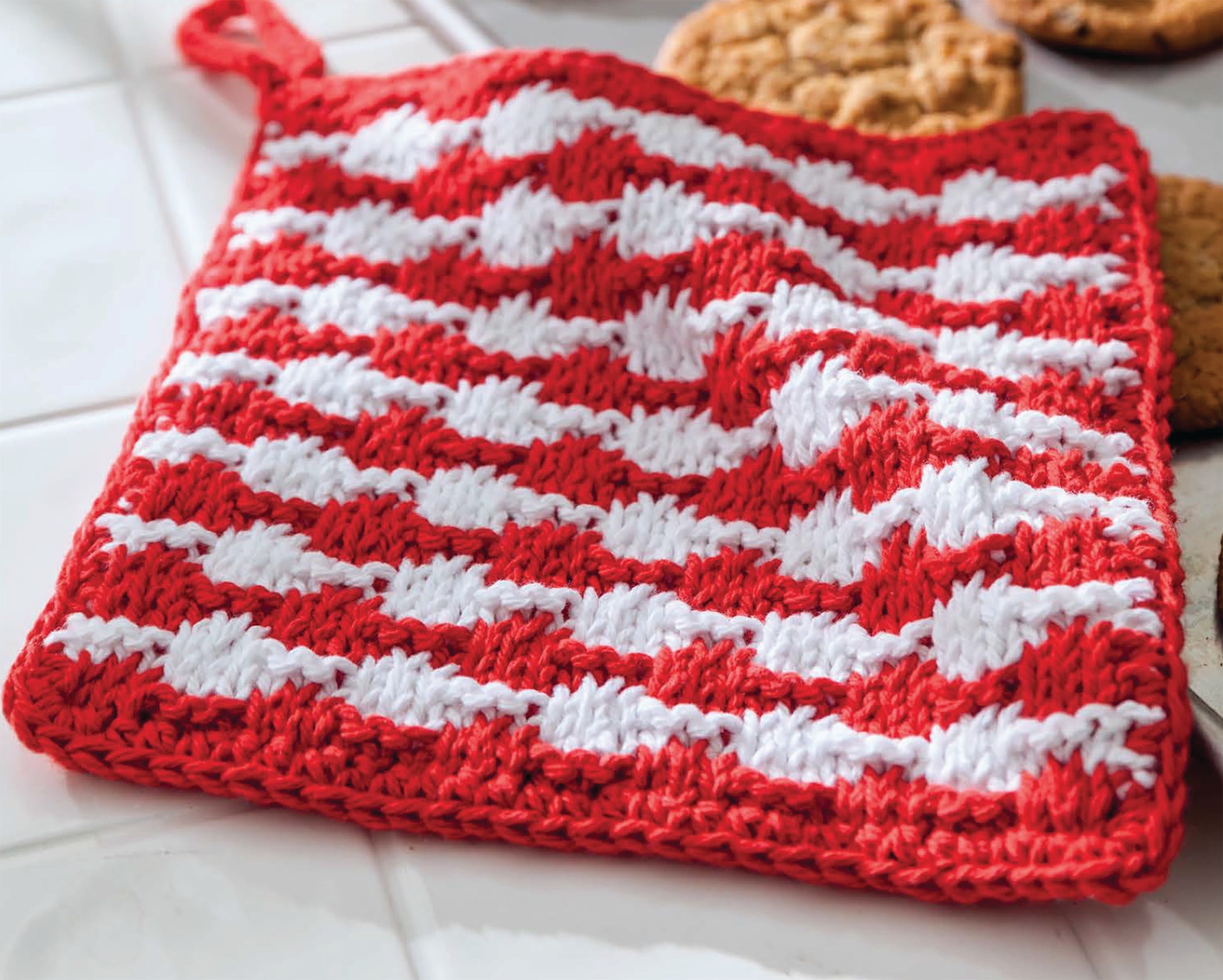 A red and white crocheted pot holder from Leisure Arts’ Make in a Weekend Potholders and Dishcloths lies on white tiles near cookies, featuring a striped, textured pattern perfect for handmade kitchen enthusiasts.