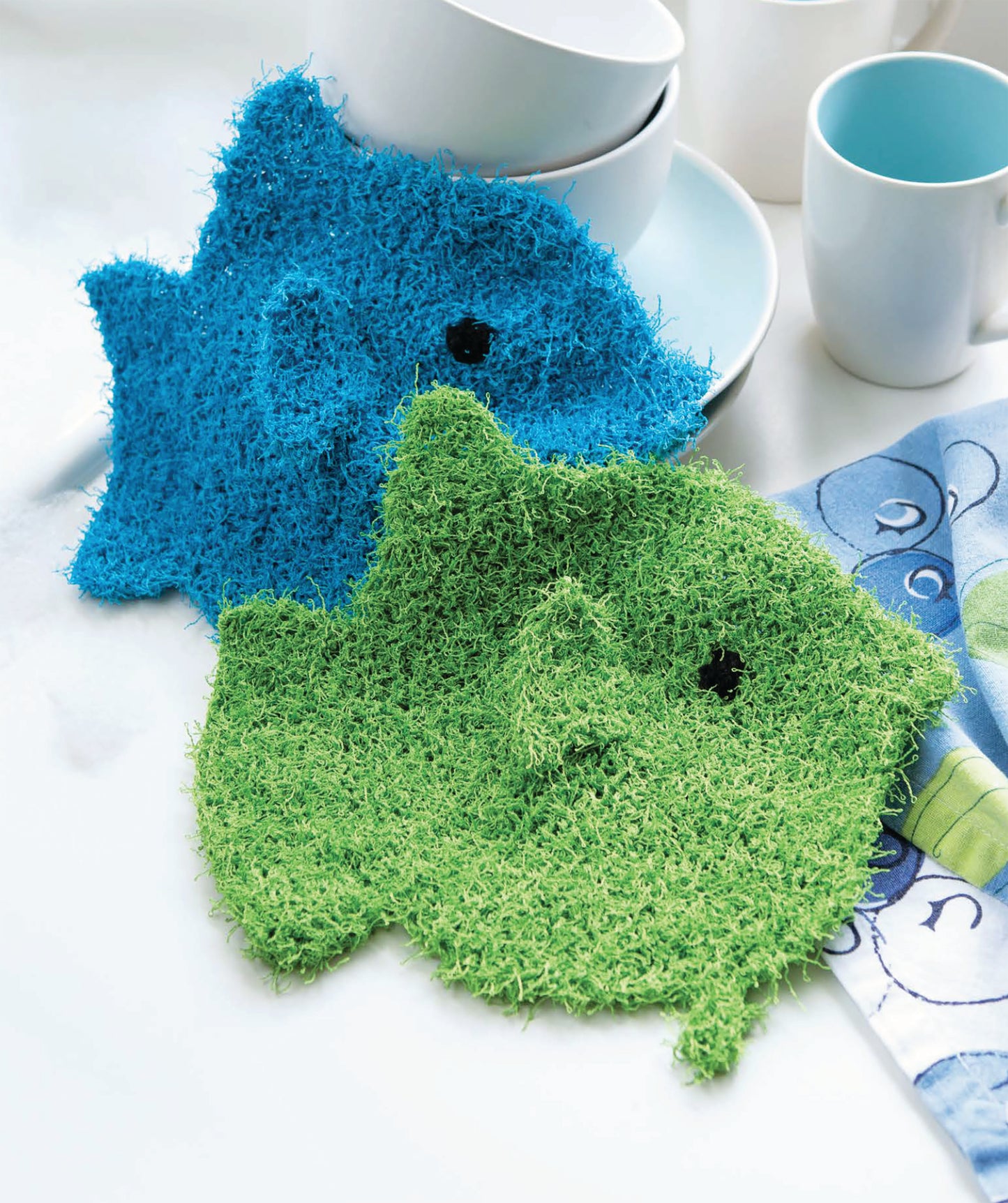Two fish-shaped dishcloths from Leisure Arts' "Make in a Weekend Potholders and Dishcloths" rest on a white countertop near stacked bowls, cups, and a blue-patterned napkin.