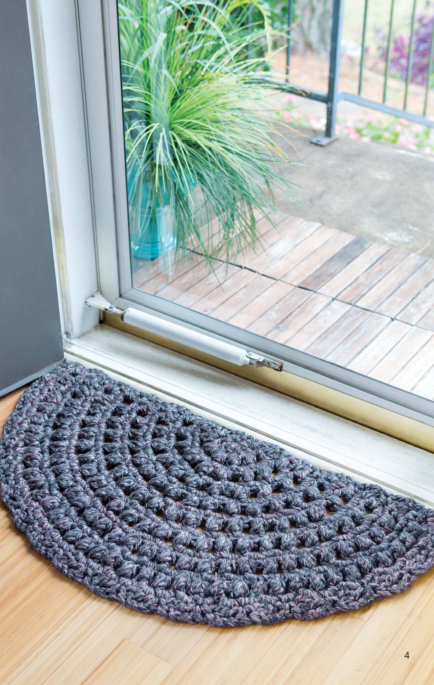 The Half Moon Rugs by Leisure Arts, crocheted in gray from beginner-friendly patterns, rest inside a glass door on a wooden floor, leading out to a patio with potted plants and garden views.