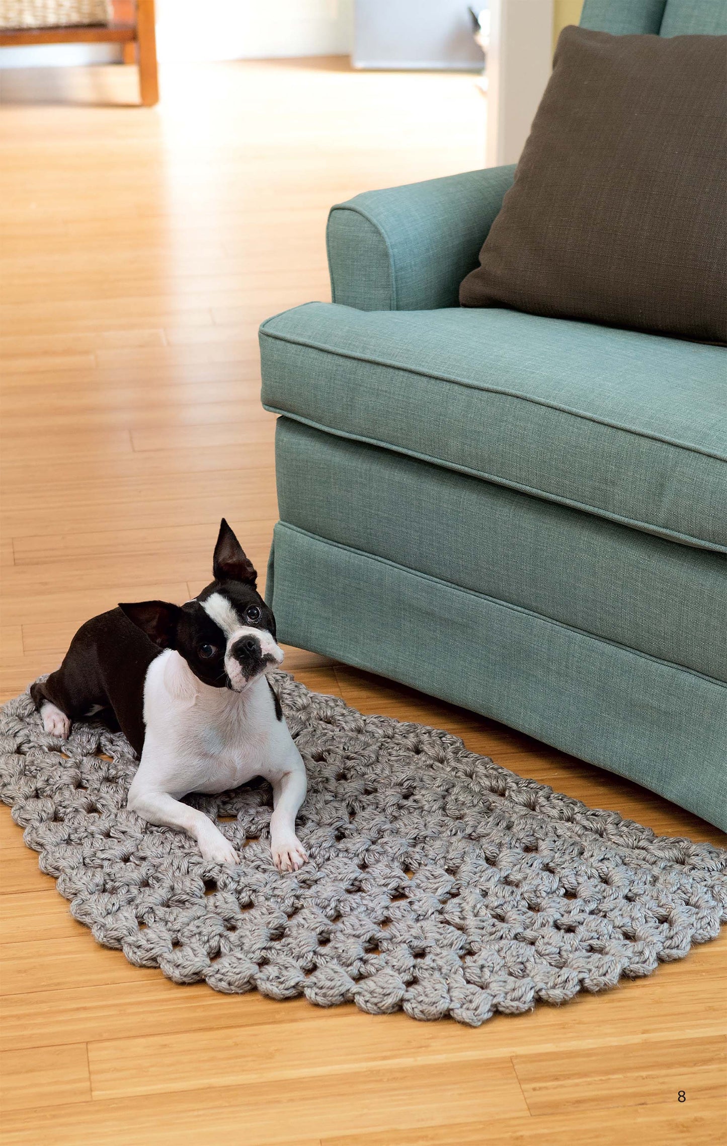 A black and white Boston Terrier lies on a chunky gray Half Moon Rug by Leisure Arts, inspired by beginner crochet patterns, next to a light teal armchair on a wooden floor in a cozy, well-lit room.