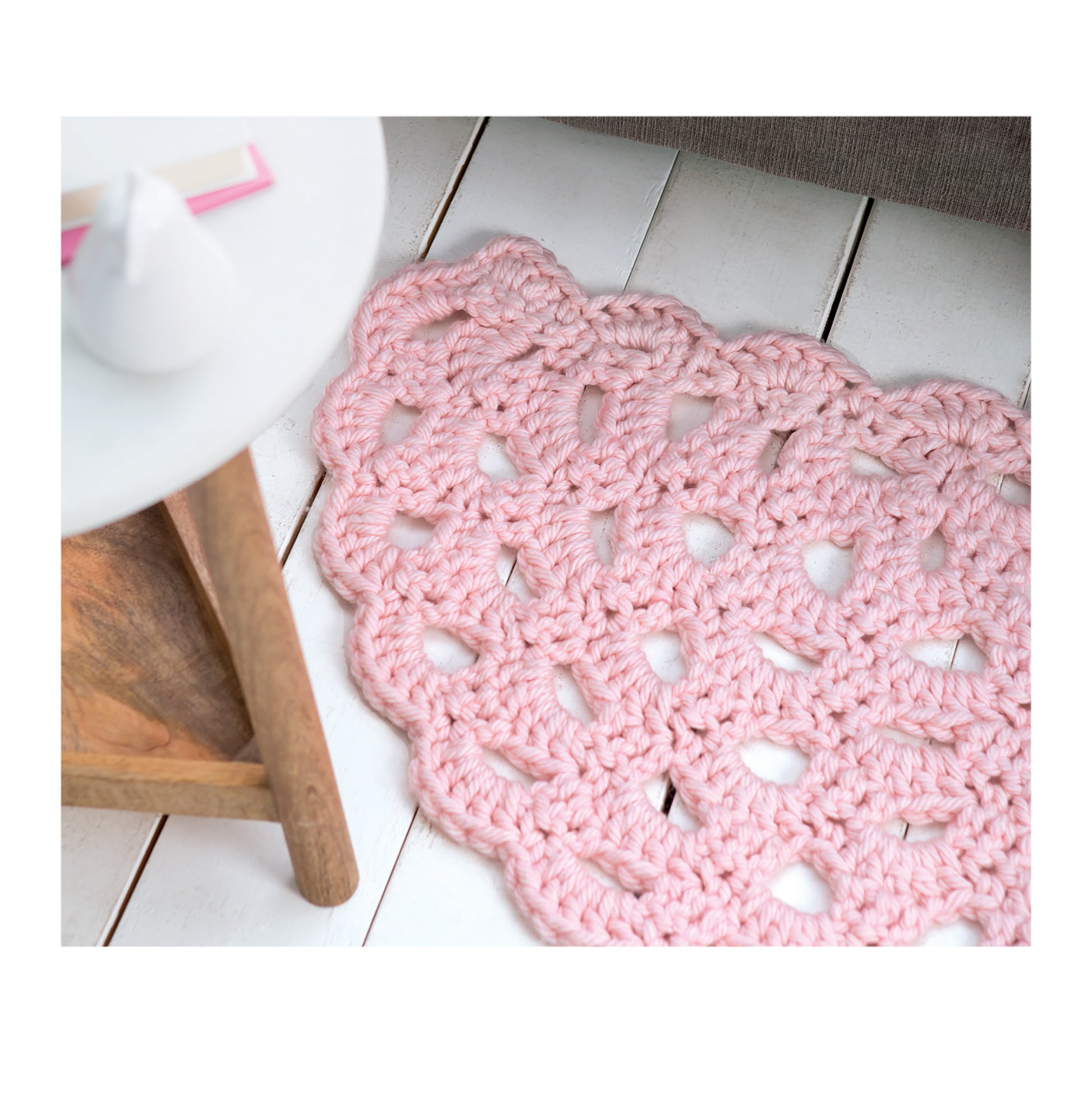 The Leisure Arts Half Moon Rug in pink features a scalloped edge and sits on white wooden floorboards by a round side table—perfect inspiration for beginner crochet projects or unique kitchen door mats.