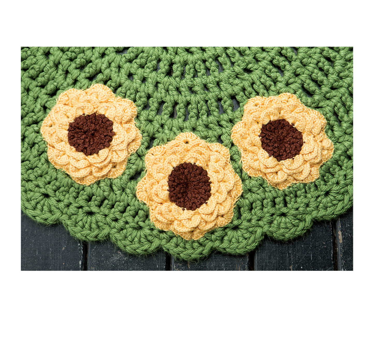 The Leisure Arts Half Moon Rugs features three yellow crocheted flowers with brown centers on a green scalloped background, creating a charming kitchen door mat for beginners. Displayed on a dark wooden surface.