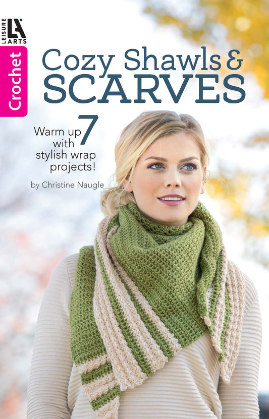 A woman in a green and beige crochet shawl stands outdoors. Text: Cozy Shawls & Scarves by Leisure Arts—7 stylish crochet patterns for shawls using medium weight yarn!.
