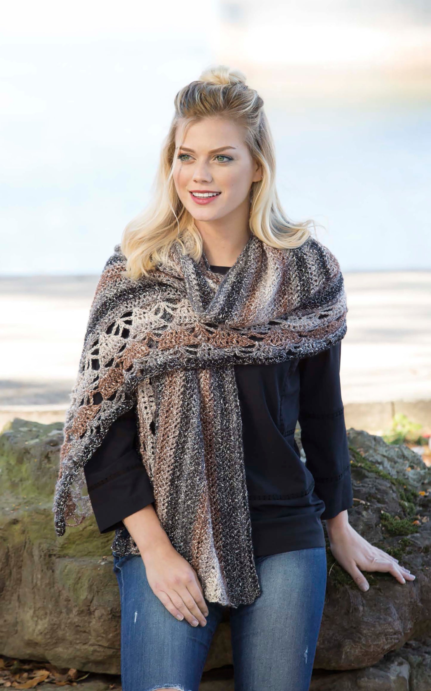 A blonde woman wears a colorful shawl made from Cozy Shawls & Scarves by Leisure Arts. Seated on a rock outdoors, she smiles and looks to the side, with trees and a blurred park in the background.