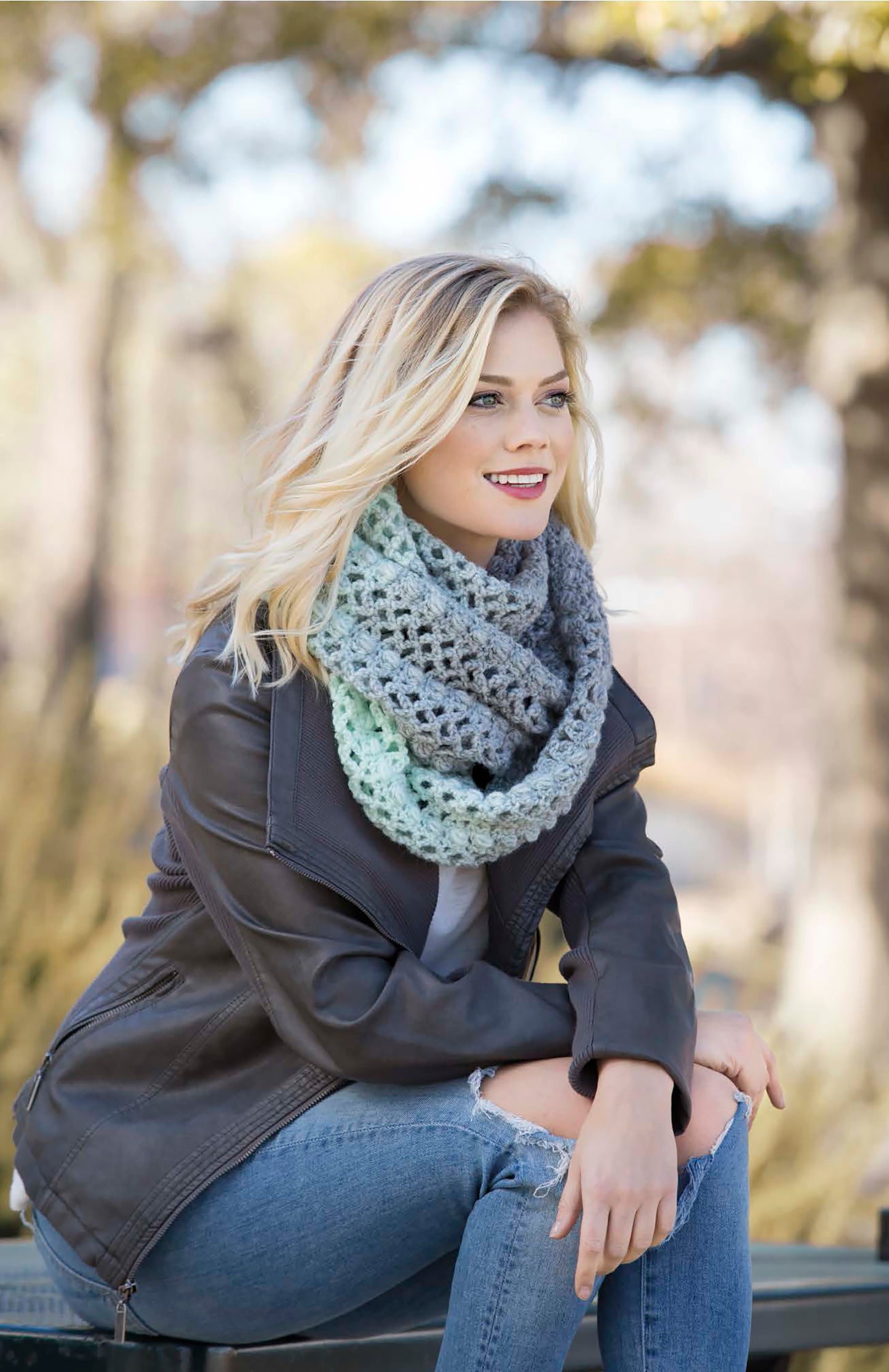 A blonde woman sits outdoors on a bench, smiling, wearing the Cozy Shawls & Scarves by Leisure Arts—a gray crochet scarf made with medium weight yarn—paired with a black leather jacket and ripped jeans; trees blur in the background.