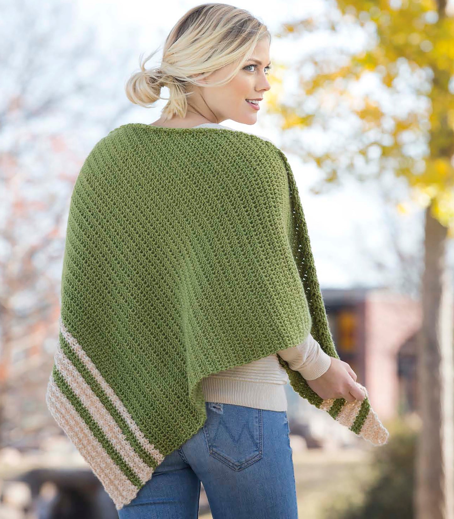 A woman with blonde hair in a loose bun models the Cozy Shawls & Scarves by Leisure Arts, featuring a green knit shawl with beige stripes made from medium weight yarn, standing outdoors amid autumn trees.