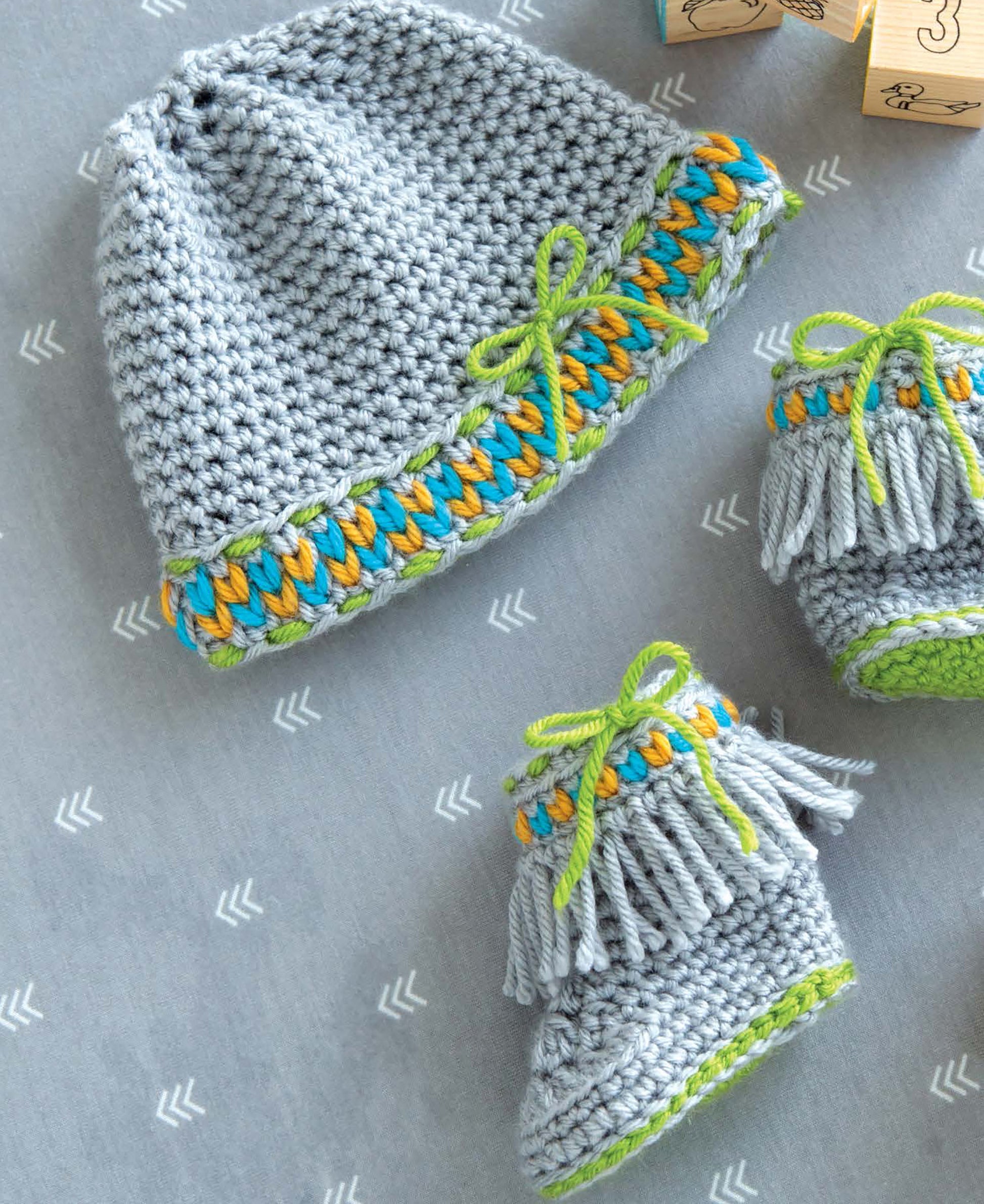 Leisure Arts Infant Boots & Hats are crocheted in gray yarn with blue, yellow, and green accents. Perfect for beginners, these modern baby accessories feature fringes and bows, shown on a gray patterned surface near wooden blocks.