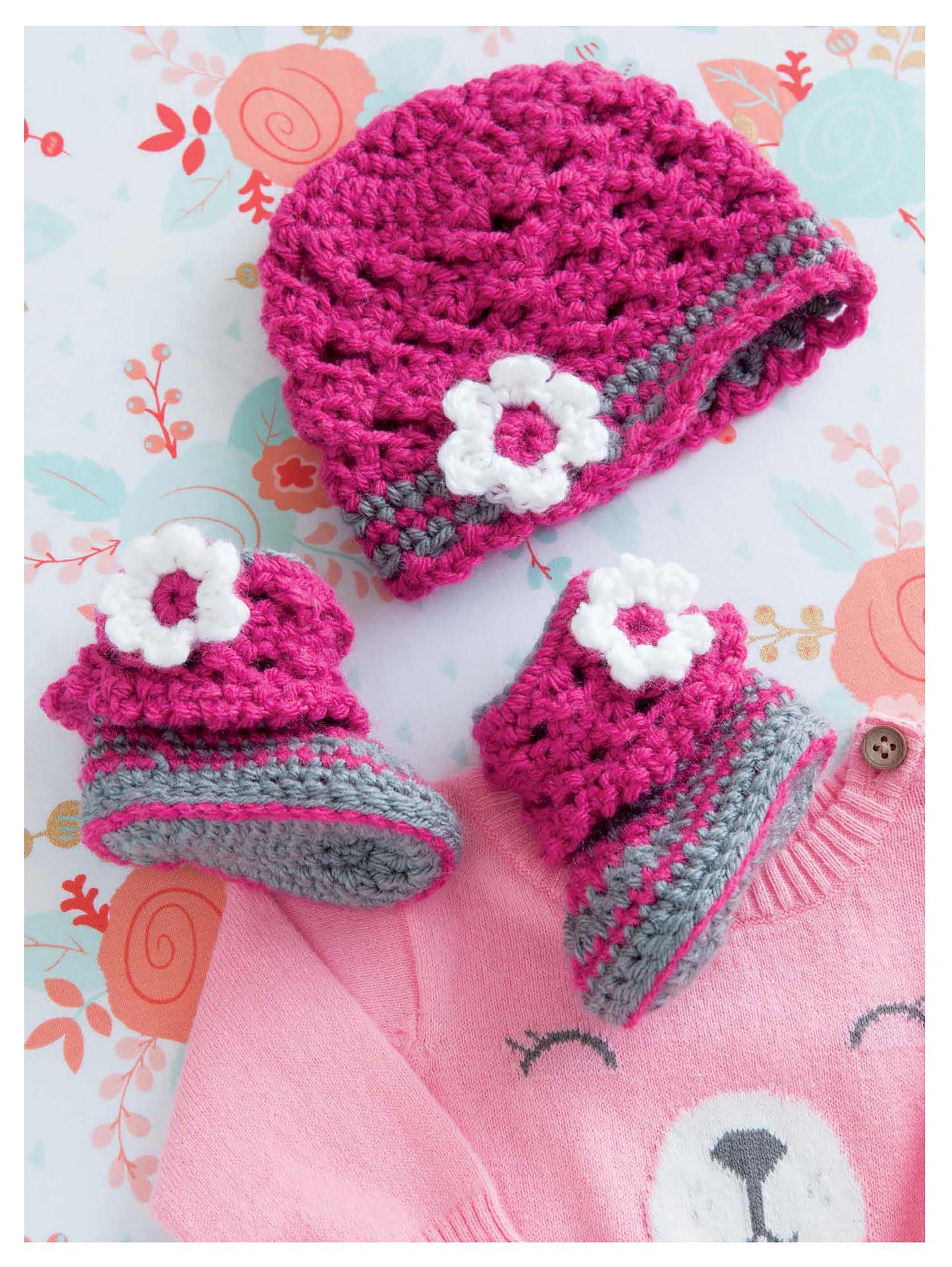 The Leisure Arts Infant Boots & Hats set, featuring a pink and gray crocheted baby hat with a white flower and matching booties, is displayed on a bear-face pink sweater atop a floral background.