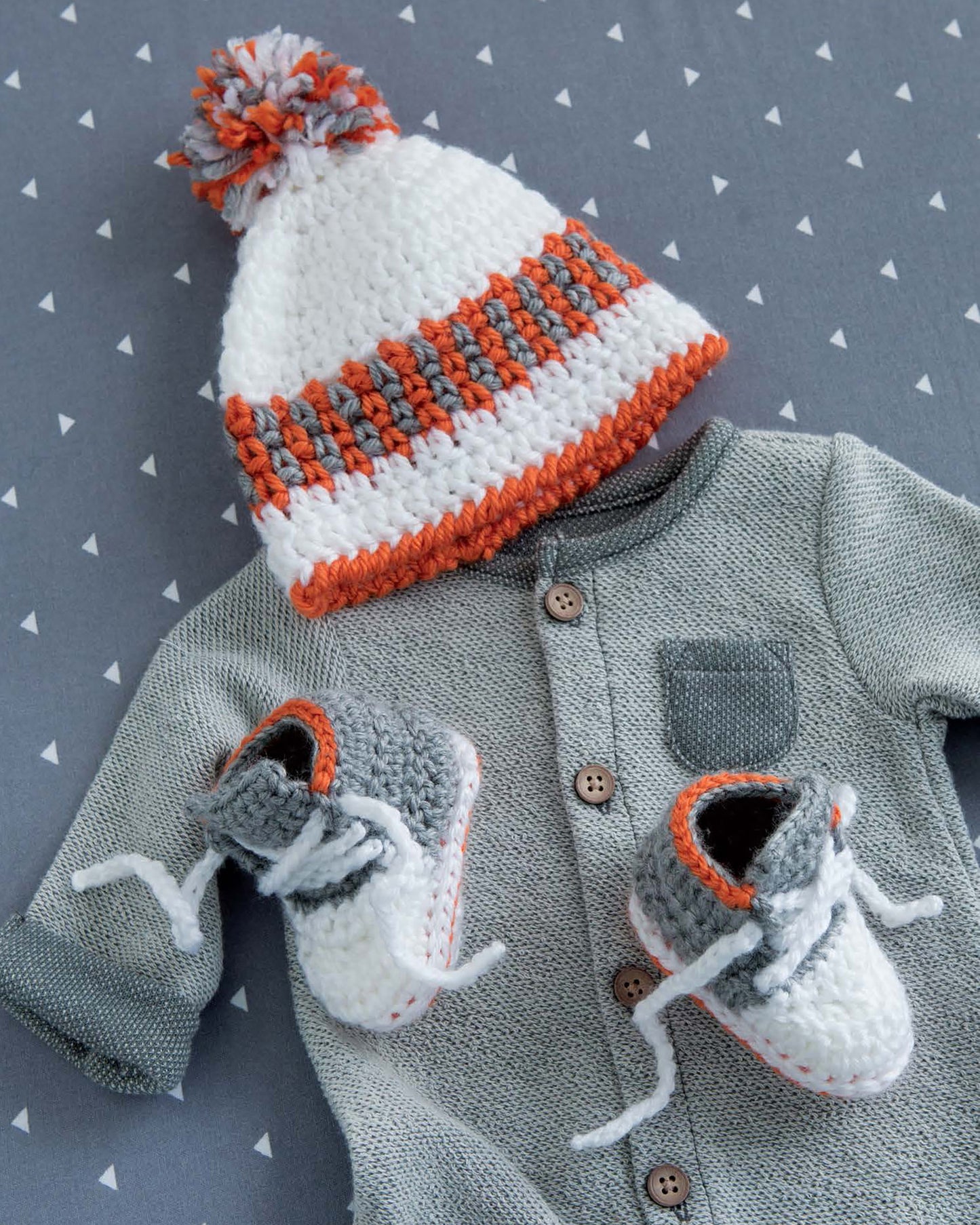 A gray baby onesie with brown buttons is displayed on a patterned surface, featuring Leisure Arts Infant Boots & Hats—a modern crocheted white, gray, and orange striped hat with matching gray and white beginner crochet booties.