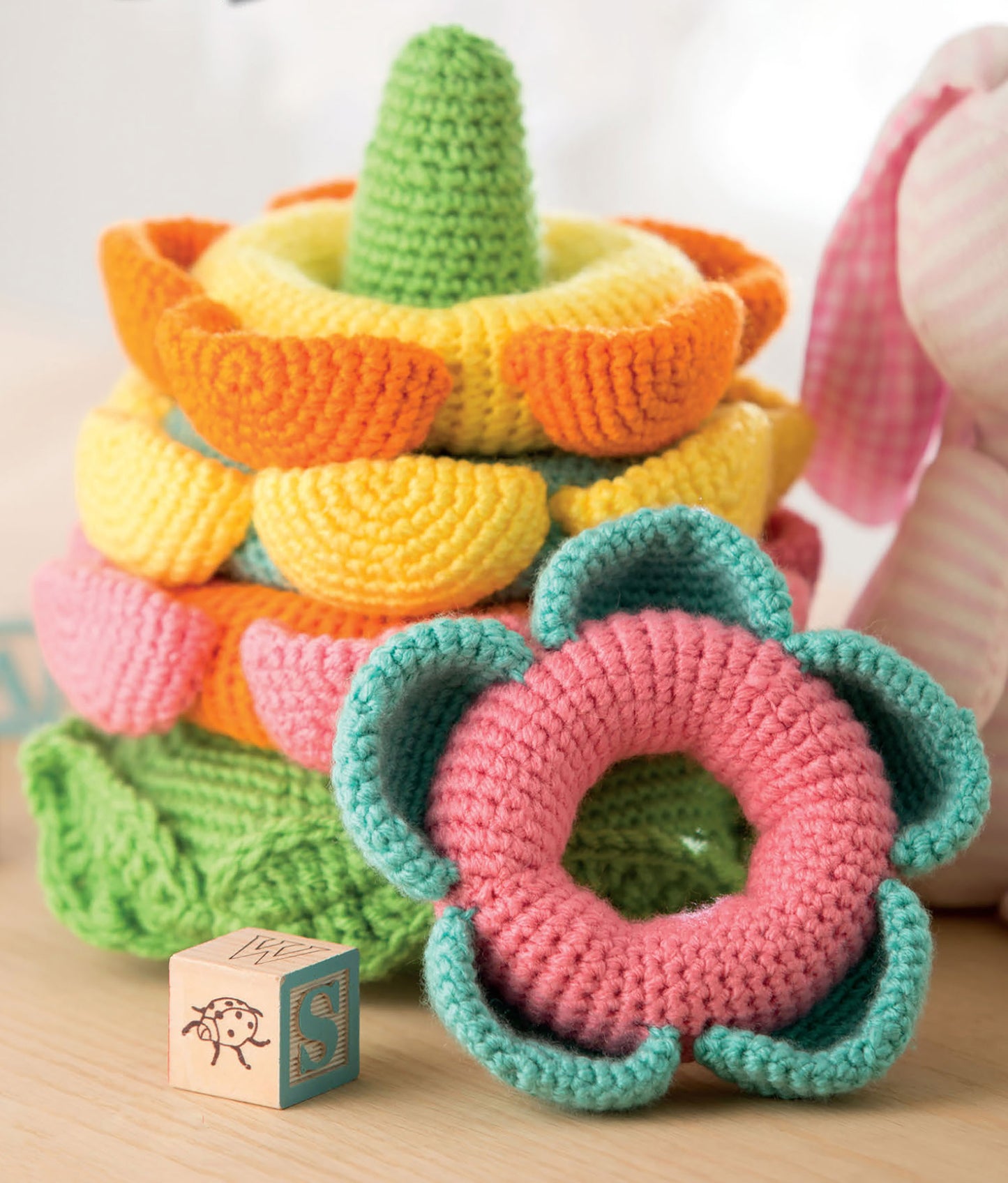 A colorful flower-shaped crochet toy from Leisure Arts' "Baby Stacks—6 Easy Toys to Crochet!" sits by a wooden alphabet block and a pink stuffed animal, highlighting charming beginner crochet designs.