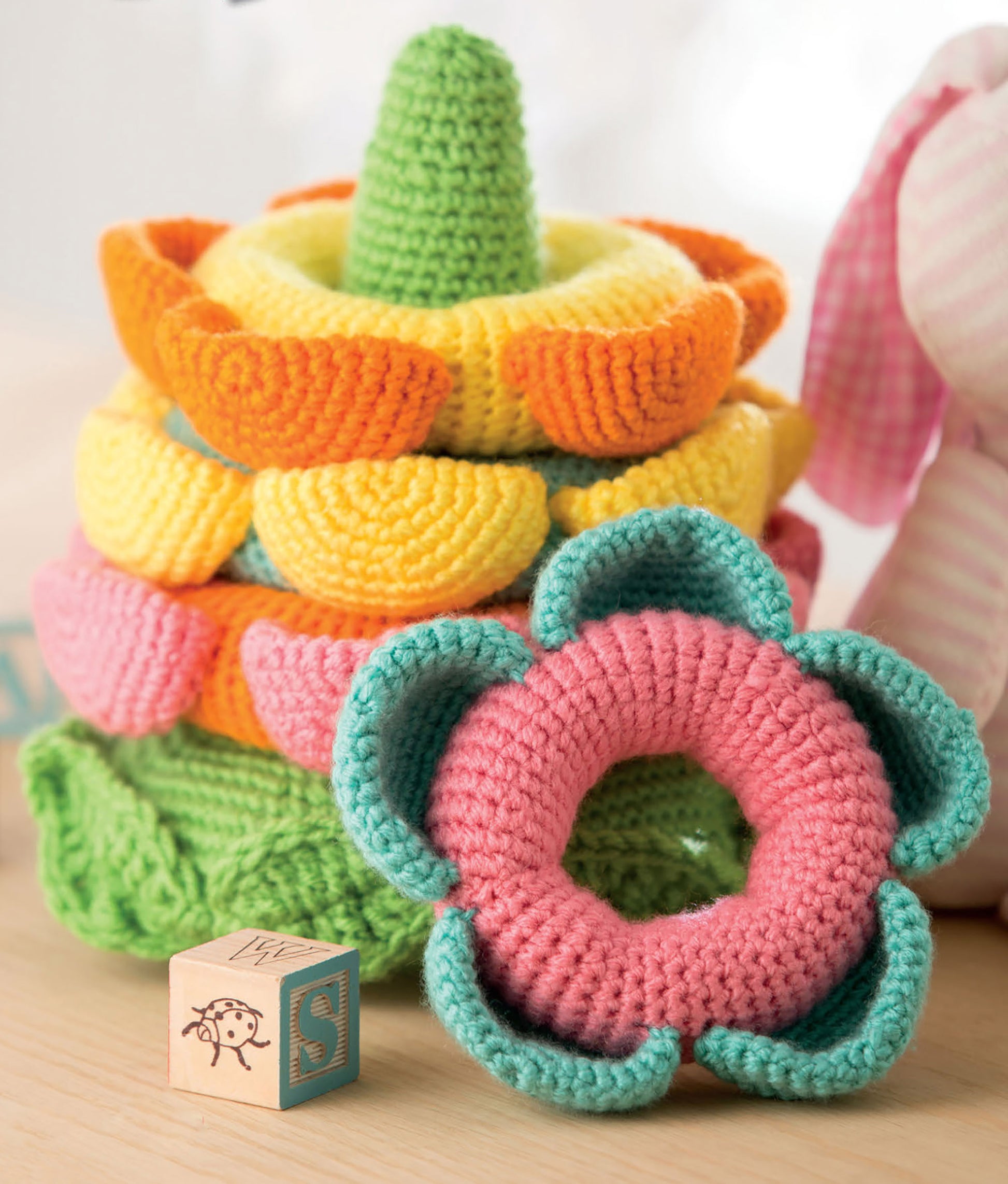 A colorful flower-shaped crochet toy from Leisure Arts' "Baby Stacks—6 Easy Toys to Crochet!" sits by a wooden alphabet block and a pink stuffed animal, highlighting charming beginner crochet designs.
