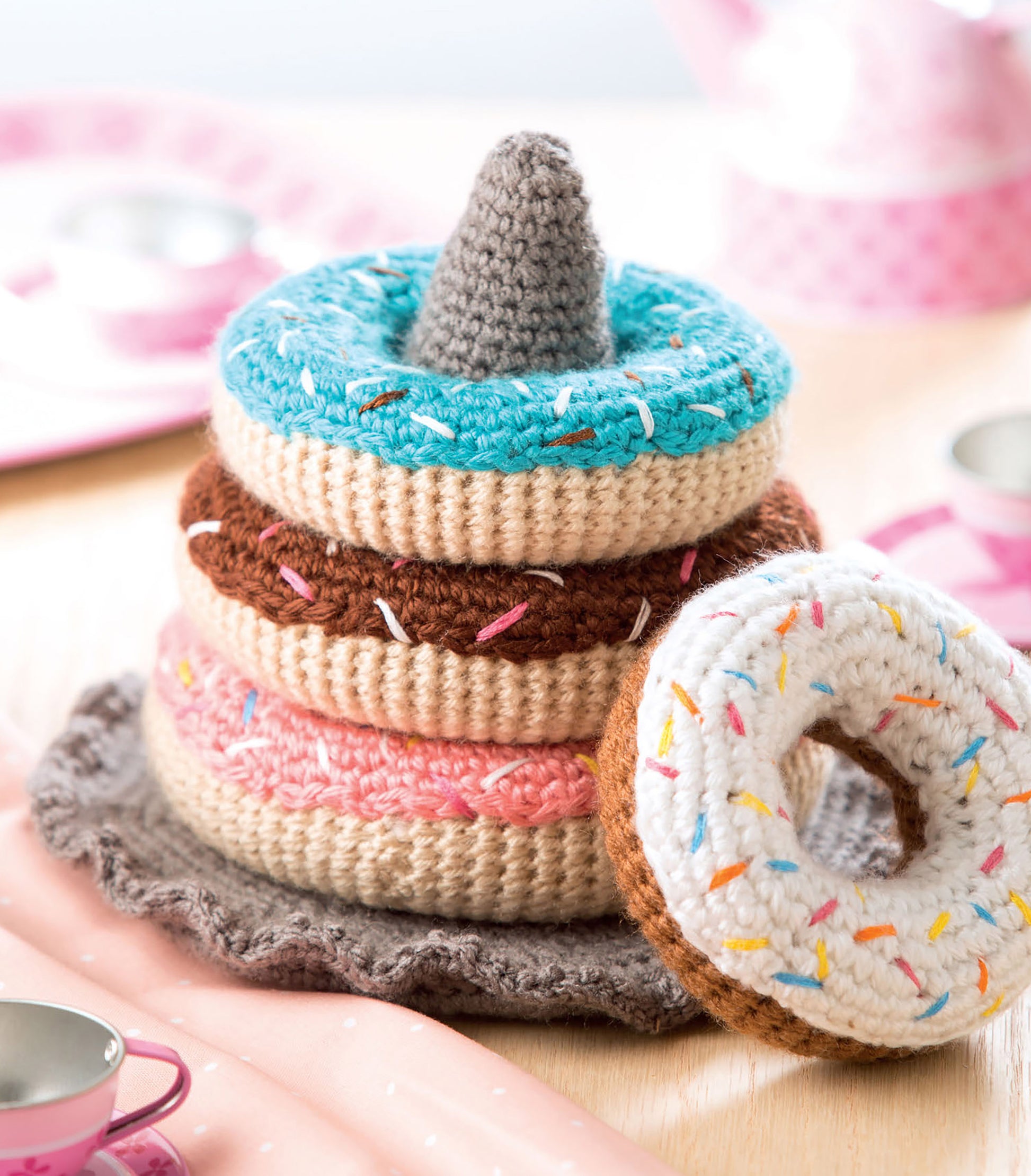A stack of colorful, crocheted toy donuts with sprinkles from "Baby Stacks—6 Easy Toys to Crochet!" by Leisure Arts sits on a soft base, while pink play dishes and cups rest in the background on a light wood surface.