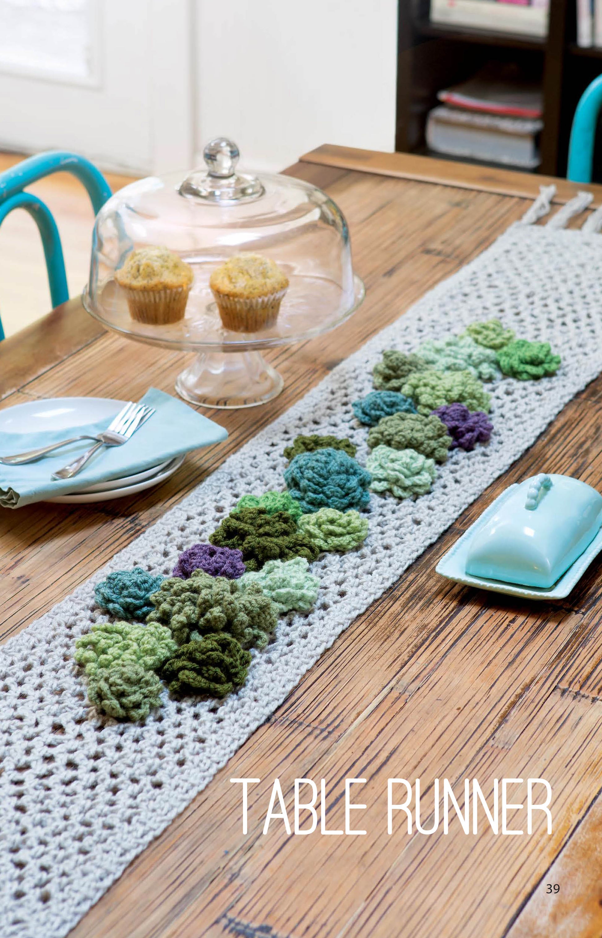 A wooden table displays the Leisure Arts Make A Crochet Garden table runner with green and purple flower motifs, a plate with silverware, a light blue butter dish, two muffins under a glass dome, and “TABLE RUNNER” at the bottom.