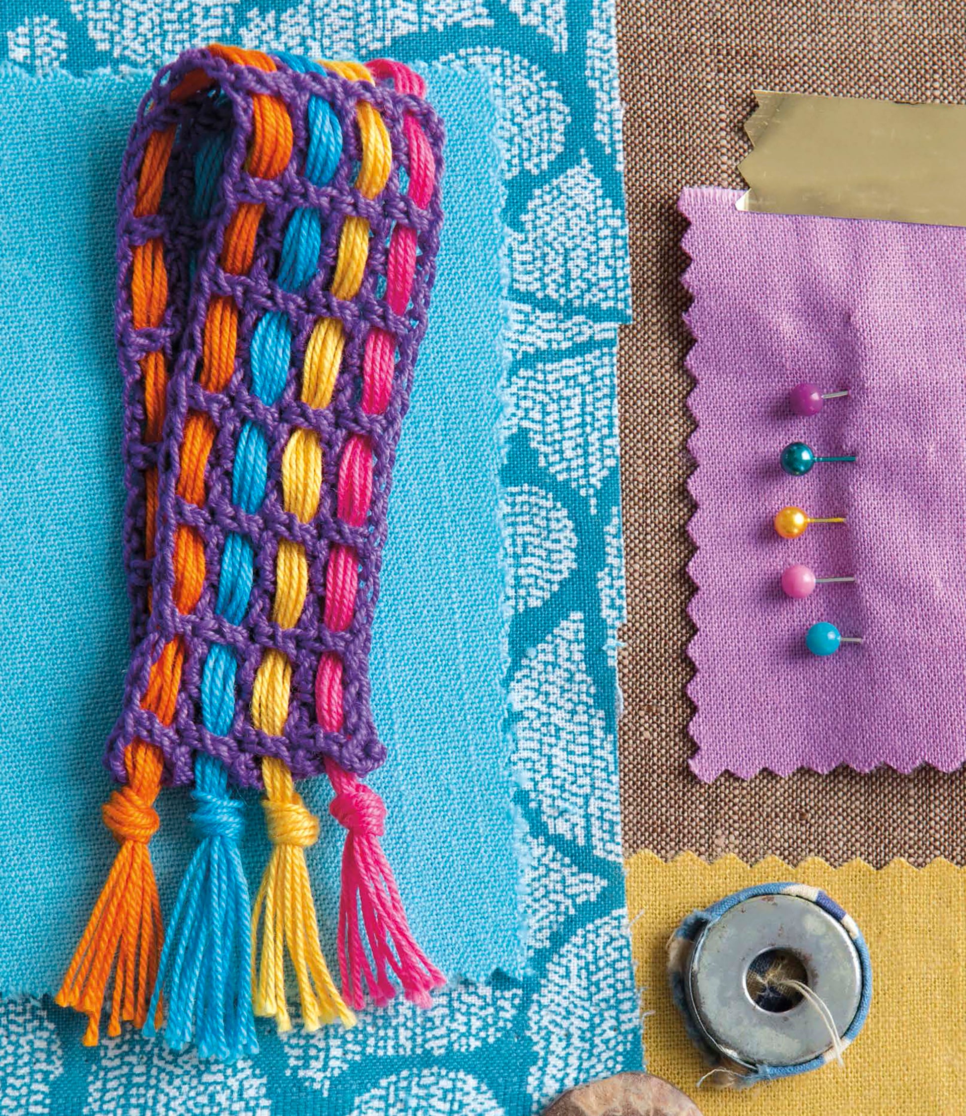 Resting on blue patterned fabric, Artisan Jewelry—9 Easy & Stylish Accents in Thread by Leisure Arts displays a woven bracelet with purple borders and rainbow threads, plus purple fabric, pins, tape, and gold foil—ideal for matching bracelets or earrings.