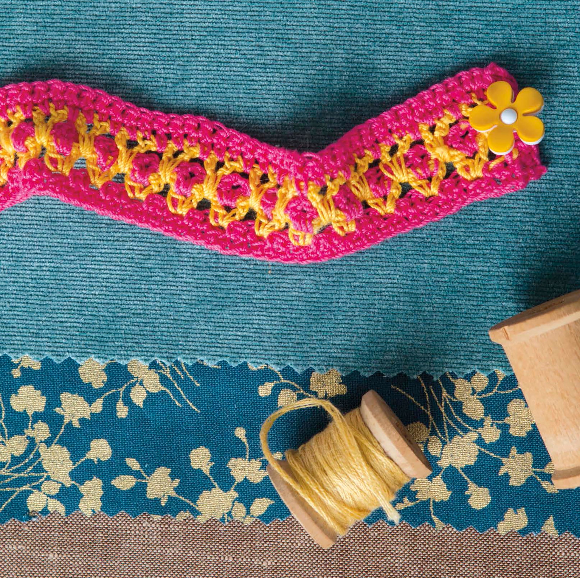A pink and yellow crocheted trim with a flower button rests on blue fabric, great for crafting jewelry like in Leisure Arts’ "Artisan Jewelry—9 Easy & Stylish Accents in Thread." Below are two wooden spools, one wound with yellow thread.