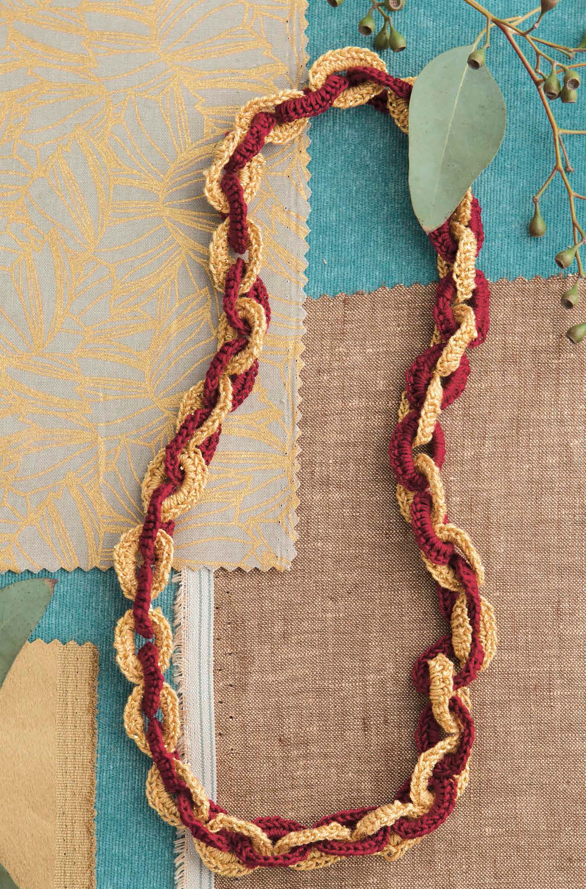 Artisan Jewelry—9 Easy & Stylish Accents in Thread from Leisure Arts features a twisted crochet necklace in gold and burgundy yarn, styled on teal, brown, and gold fabric swatches with eucalyptus and dried berries—ideal with your favorite accessories.