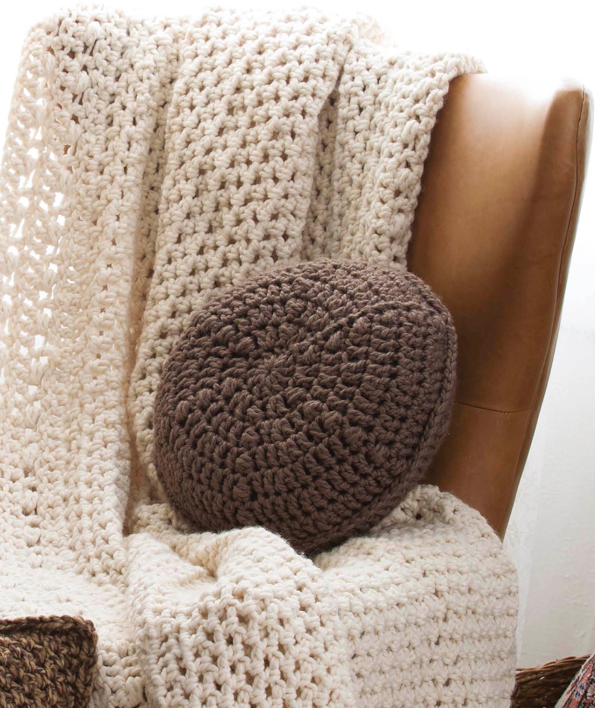 A beige crocheted blanket and a dark brown pillow on a tan leather chair showcase projects from Leisure Arts’ “Quick Crochet Home Decor”—your go-to for easy patterns, illustrated stitch guides, and step-by-step instructions.