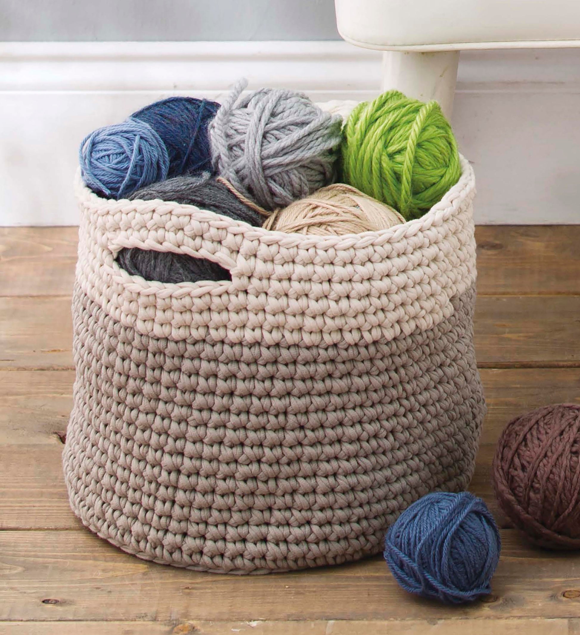 A woven basket of colorful yarn sits on a wooden floor by a white chair—perfect for creating easy projects from Quick Crochet Home Decor by Leisure Arts, featuring illustrated stitch guides and step-by-step instructions. Some yarn balls rest beside the basket.