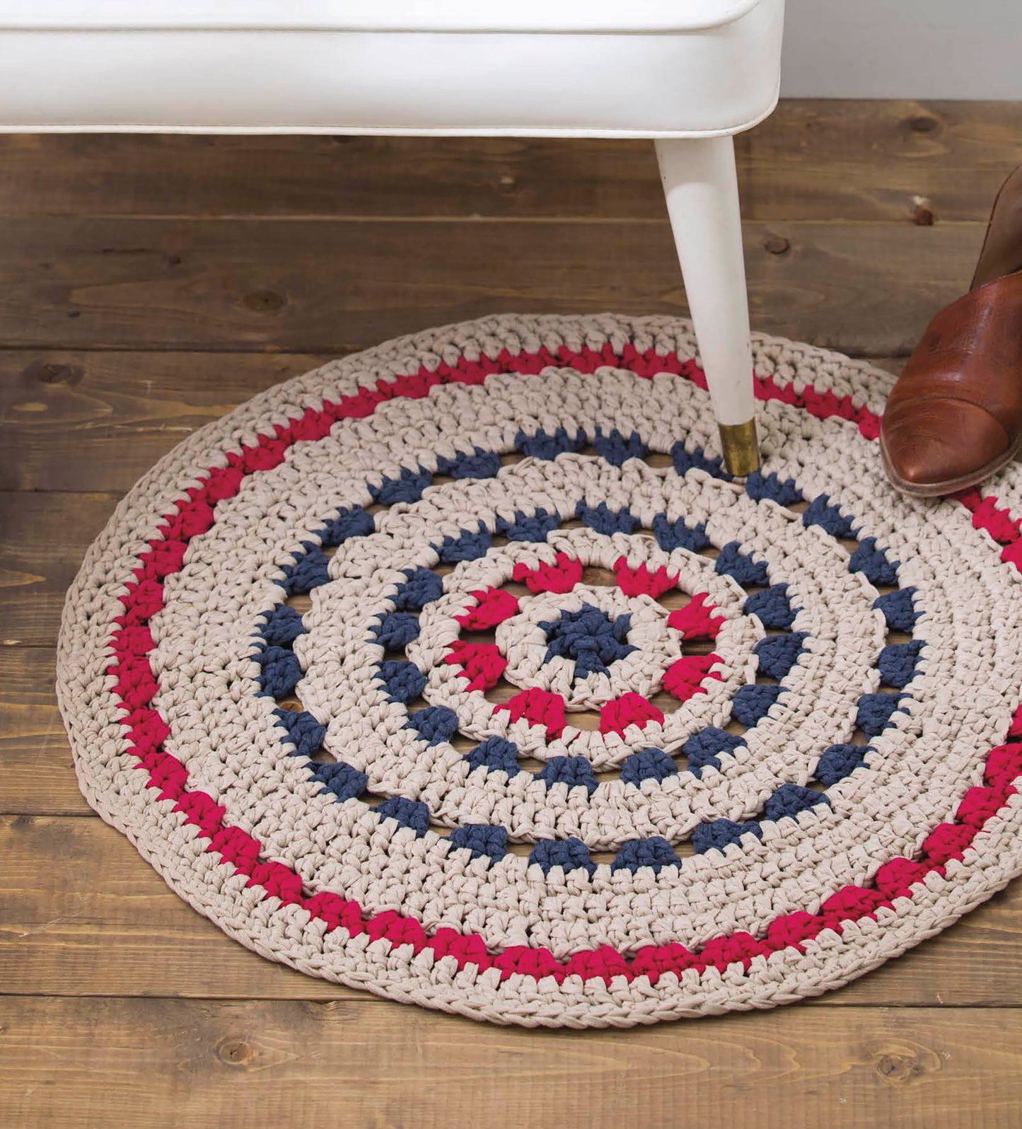 A round crocheted rug in beige, red, and navy blue stripes from "Quick Crochet Home Decor" by Leisure Arts rests on a wooden floor beside a white-legged furniture piece and a brown leather shoe—ideal inspiration for easy crochet projects.