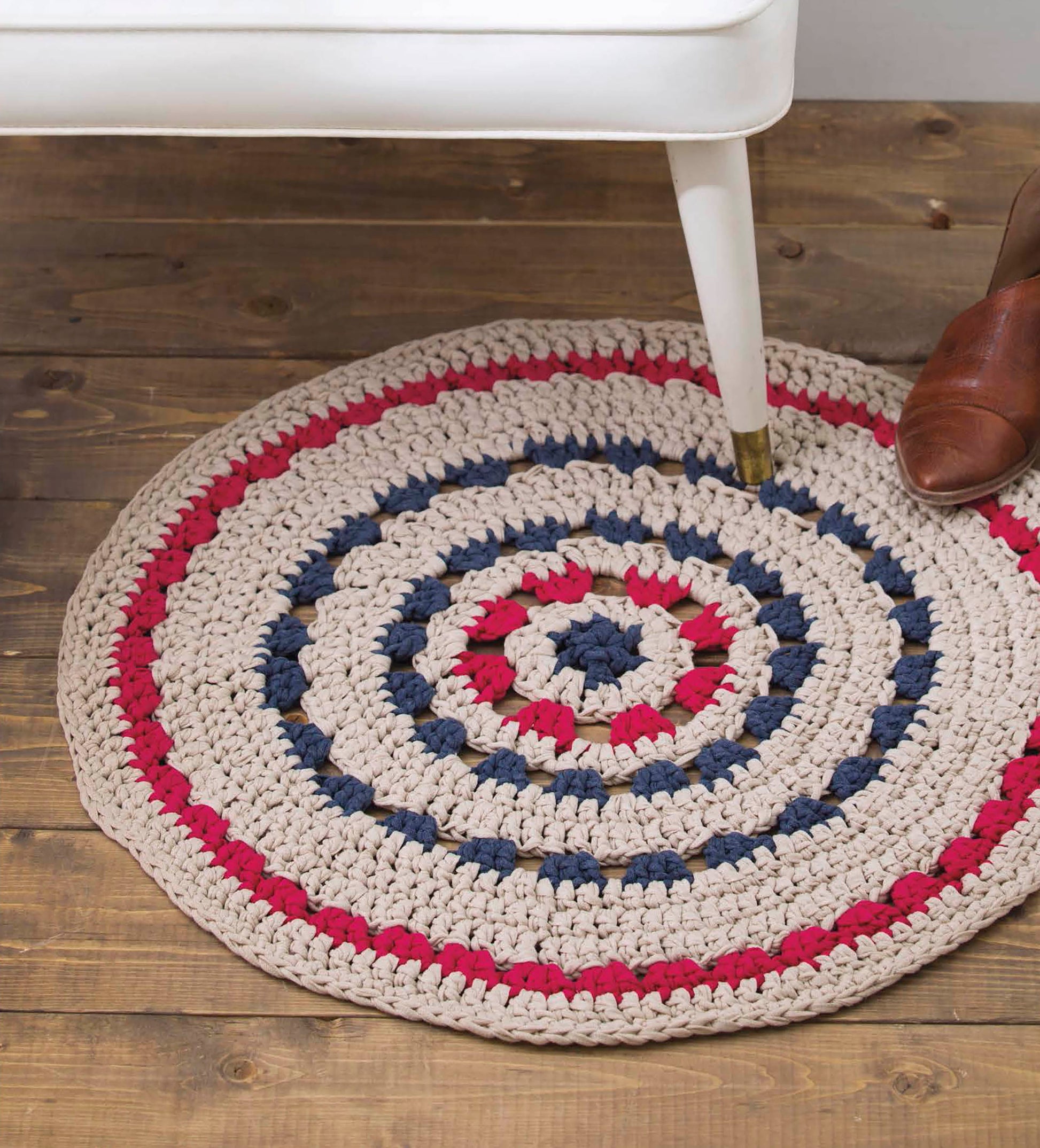 A round crocheted rug in beige, red, and navy blue stripes from "Quick Crochet Home Decor" by Leisure Arts rests on a wooden floor beside a white-legged furniture piece and a brown leather shoe—ideal inspiration for easy crochet projects.