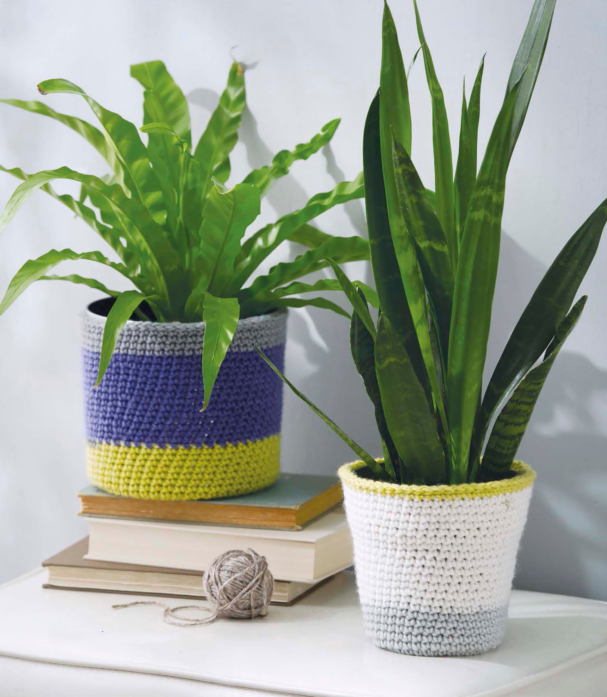 Two potted houseplants on books and a white table have pots covered in easy crochet cozies from Quick Crochet Home Decor by Leisure Arts. A ball of gray yarn nearby invites creativity and simple crochet projects for your home.