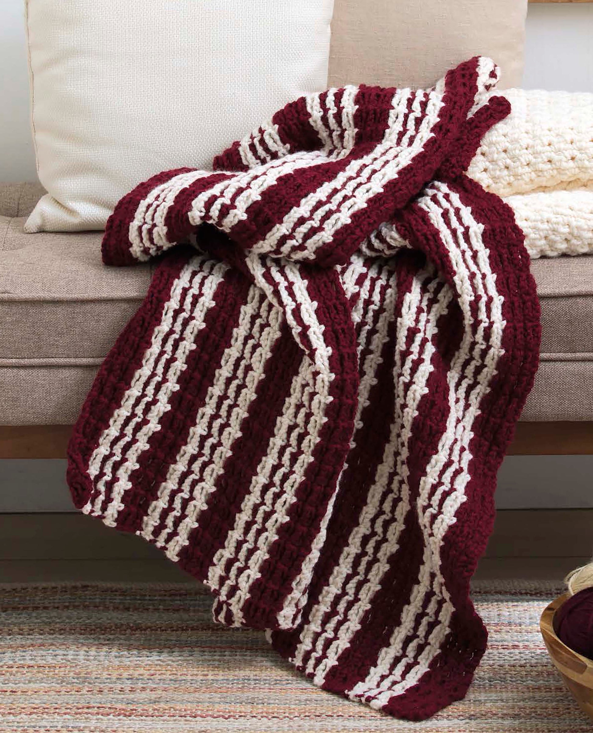 A crocheted blanket made from Leisure Arts' "Make in a Weekend Textured Lap Throws Digital Download" with wide maroon and cream stripes drapes over a beige sofa accented by a white pillow, textured cream blanket, and a woven rug on the floor.