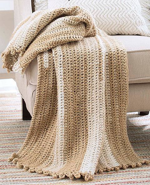 The Make in a Weekend Textured Lap Throws Digital Download by Leisure Arts features a beige and cream striped crocheted blanket, shown draped over a light armchair with a patterned pillow and woven rug in the background.