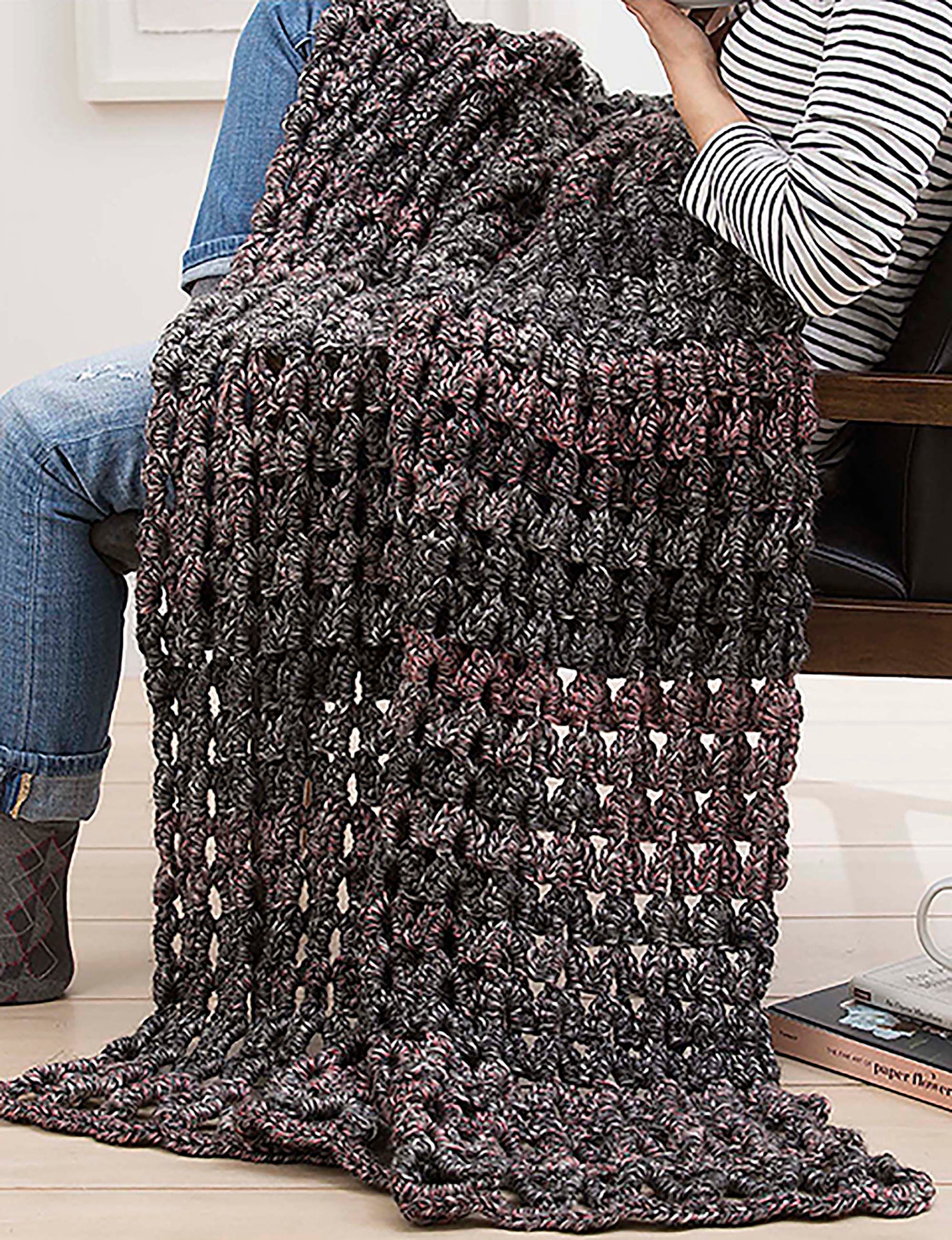 A person sits with a Make in a Weekend Textured Lap Throw by Leisure Arts, featuring a loose, open stitch pattern in gray and dark purple, draped over their lap—ideal for beginner or weekend crochet projects (digital download).