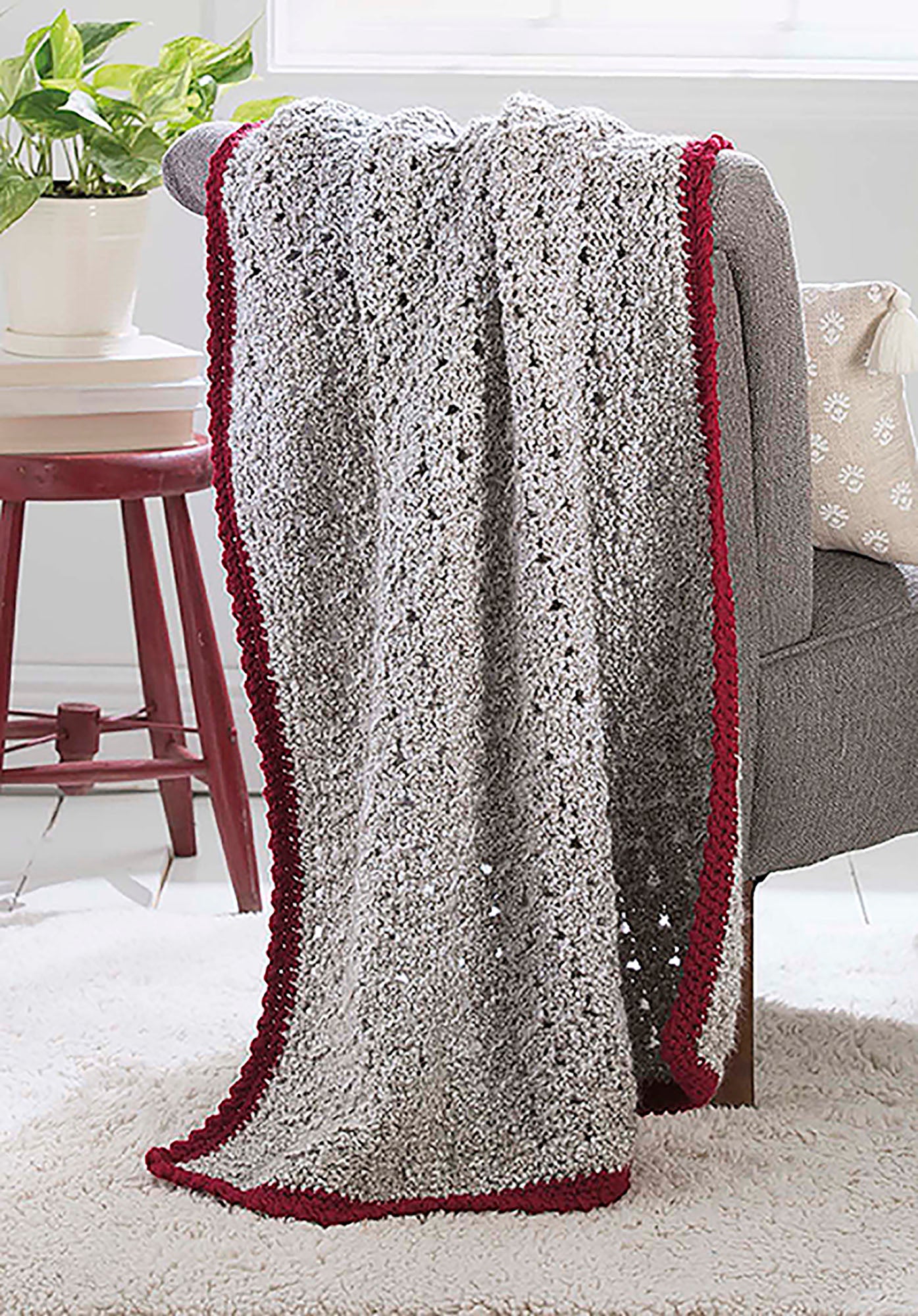 The Make in a Weekend Textured Lap Throws Digital Download by Leisure Arts features a gray crocheted blanket with a red scalloped border, shown on an armchair next to a red table and potted plant atop a white rug in a bright room.