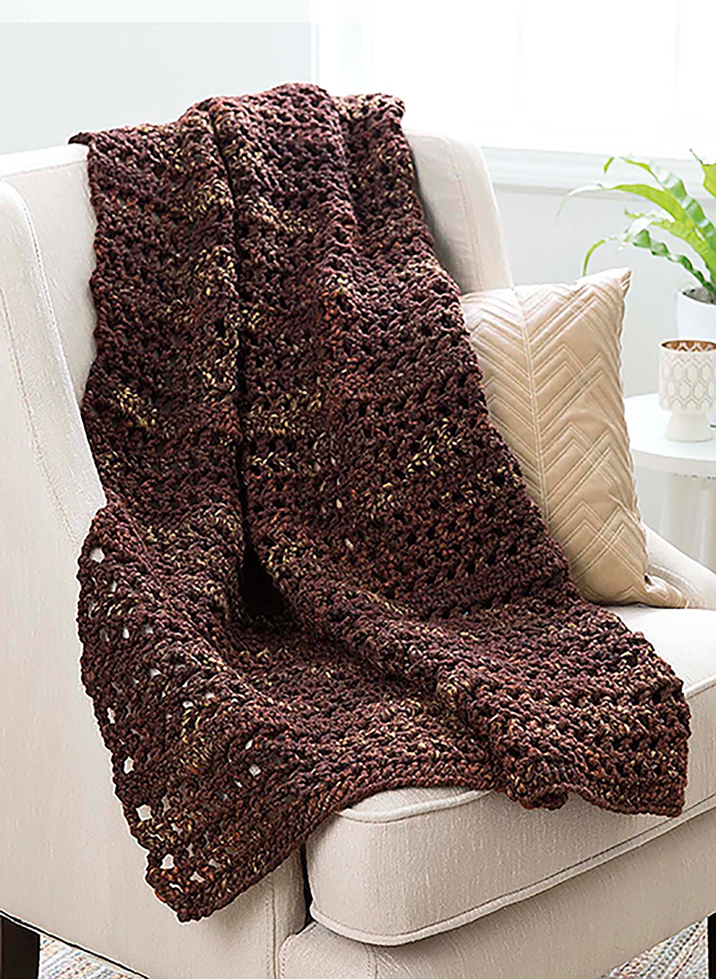 The Make in a Weekend Textured Lap Throws Digital Download by Leisure Arts features a chunky brown crochet blanket with subtle flecks, draped over a light armchair beside a textured cream cushion and a white mug on a small table.
