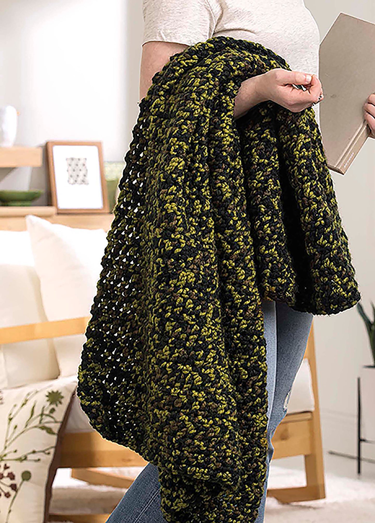 Someone holds a thick green and black blanket from Leisure Arts' "Make in a Weekend Textured Lap Throws" digital download, draped over their arm, standing indoors with a book. A cozy modern living room with white couch and wood furniture is behind them.