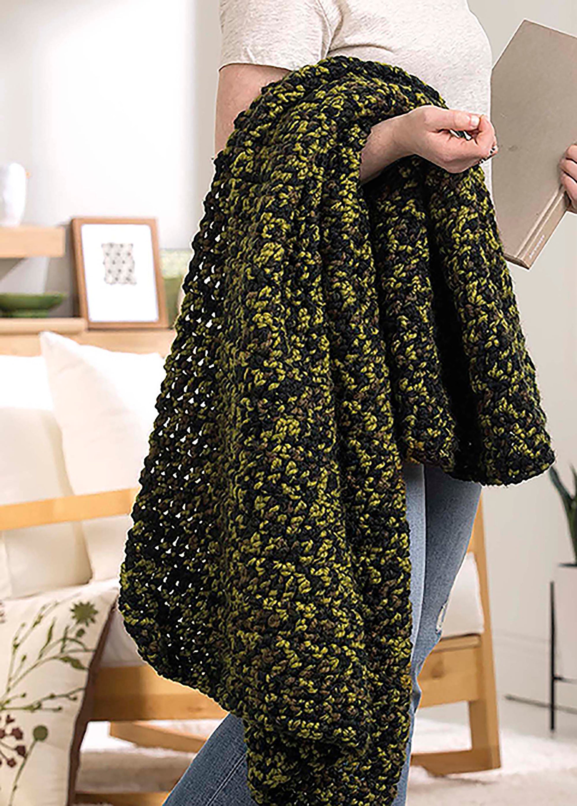 Someone holds a thick green and black blanket from Leisure Arts' "Make in a Weekend Textured Lap Throws" digital download, draped over their arm, standing indoors with a book. A cozy modern living room with white couch and wood furniture is behind them.