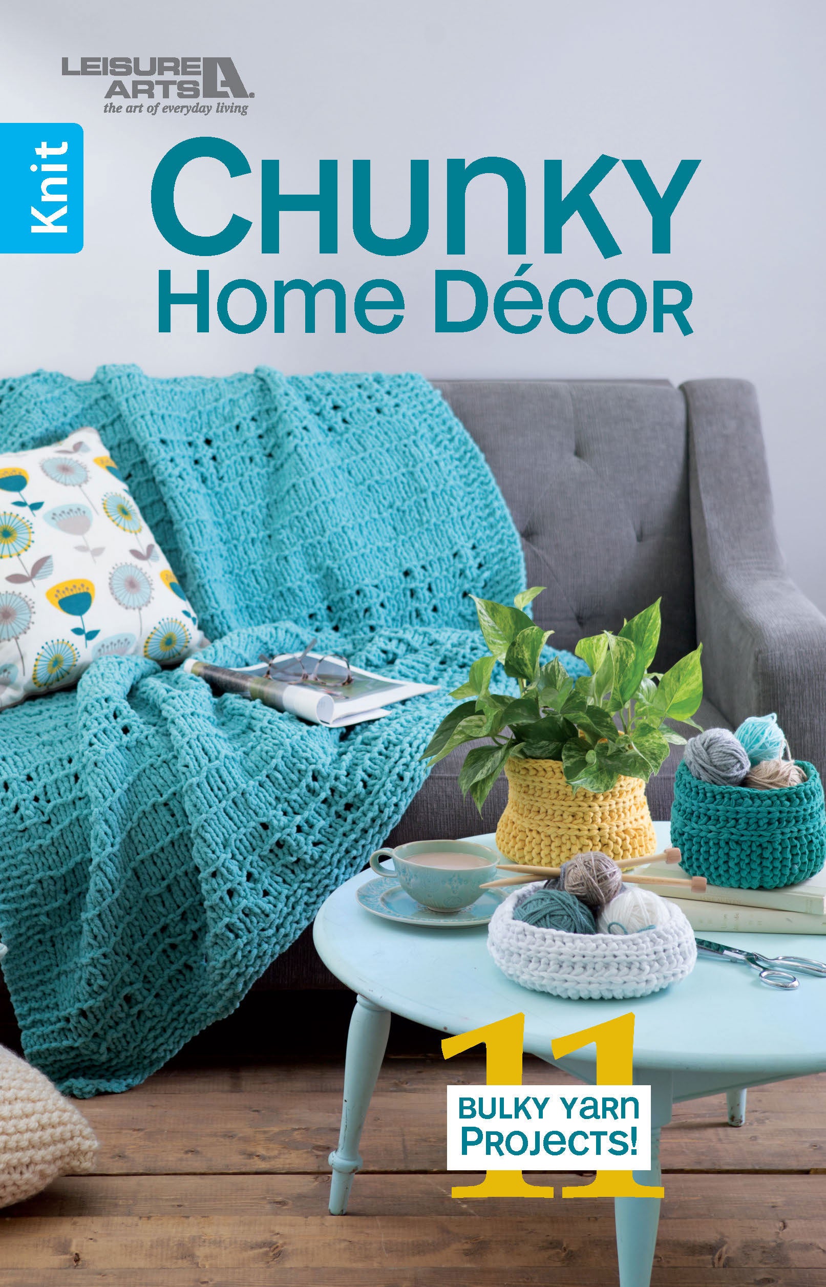 A gray sofa with a teal chunky knit blanket and floral pillow sits behind a light blue coffee table with a plant, books, and yarn baskets. Text: “Chunky Home Decor Digital Download by Leisure Arts – 11 Bulky Yarn Projects!”.