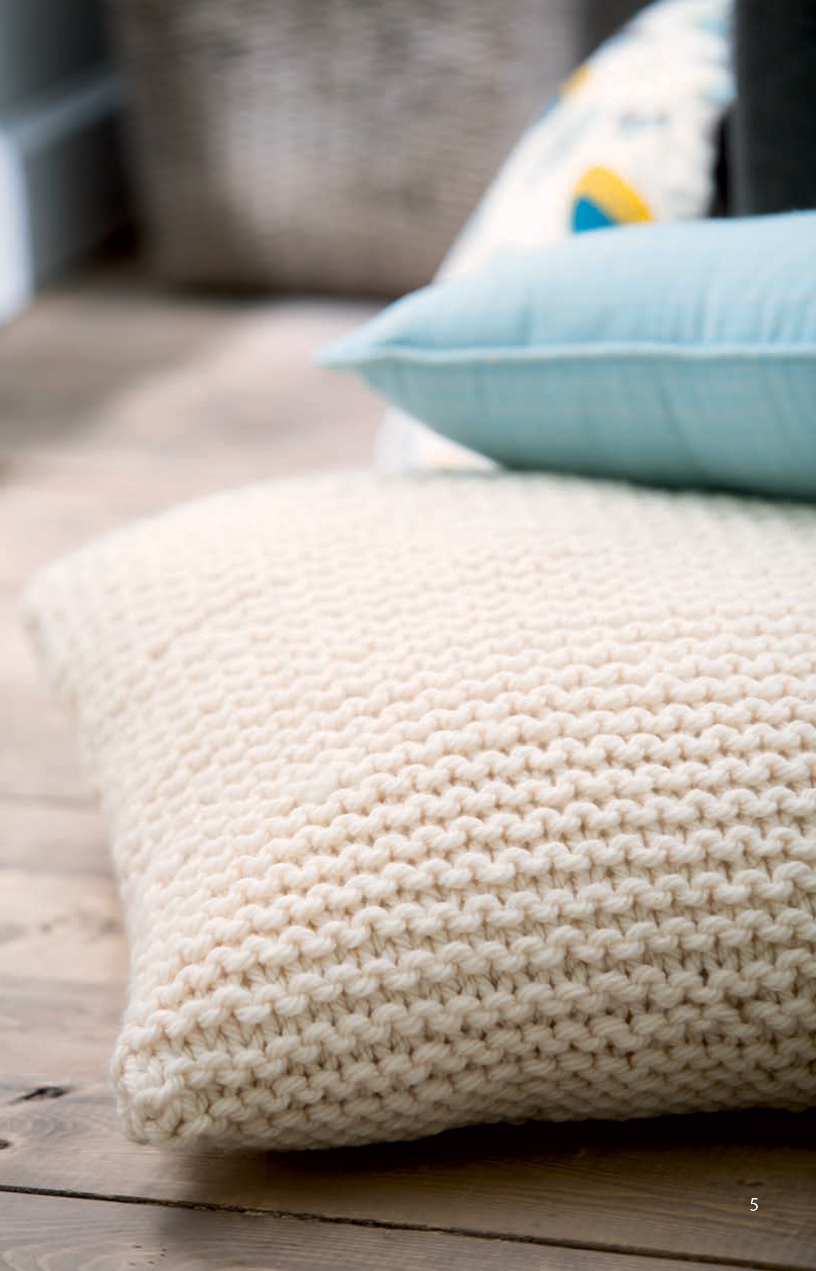 A close-up of a large cream cushion on a wooden floor, topped with layered cushions including light blue, showcases the cozy charm of the Chunky Home Decor Digital Download by Leisure Arts; a woven basket is softly blurred in the background.