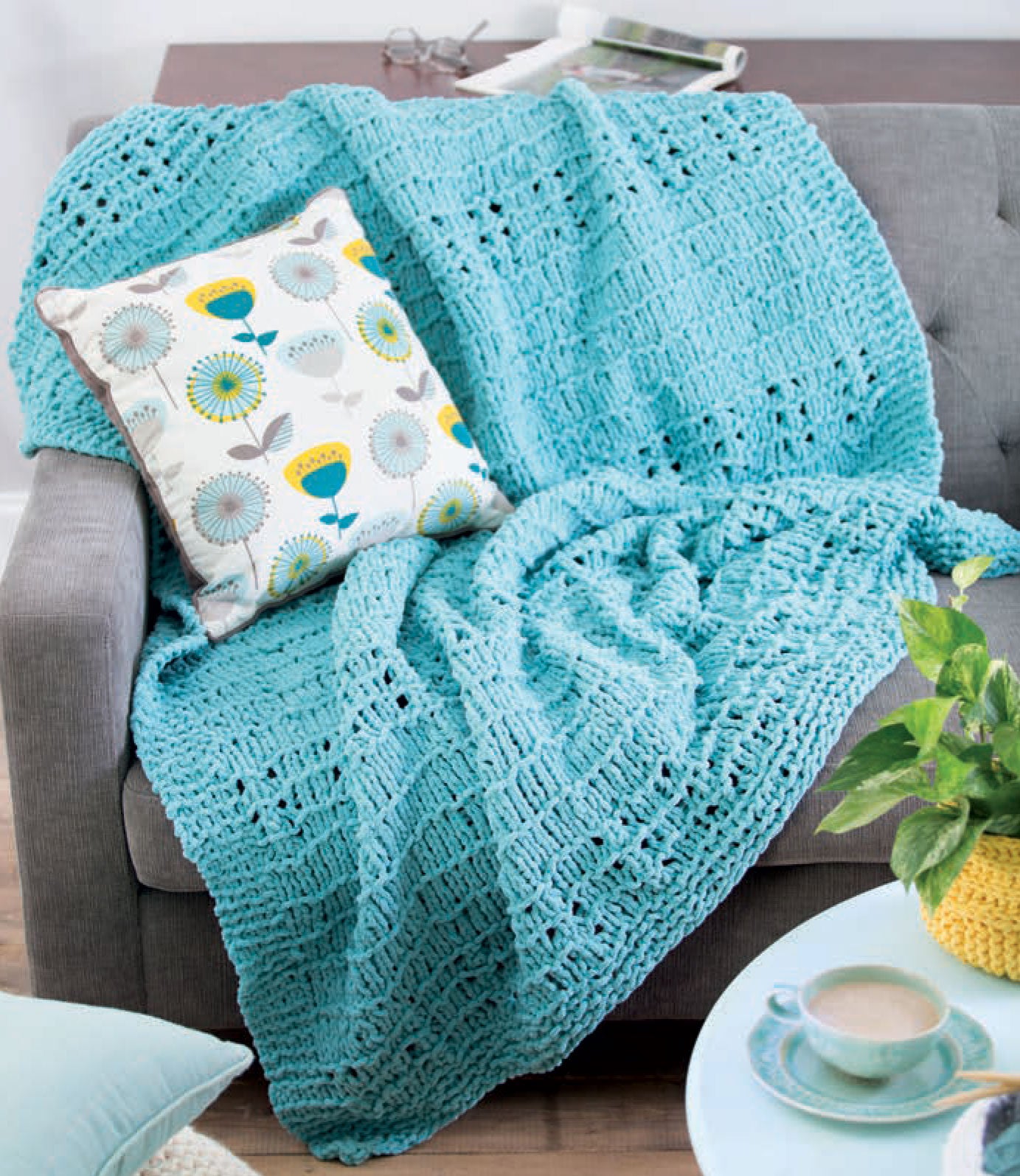 The "Chunky Home Decor Digital Download" by Leisure Arts features a cozy blue crocheted blanket, ideal for bulky yarn projects, draped over a gray sofa with floral pillow, beside a small plant & coffee cup on a blue table. Glasses and book rest nearby.