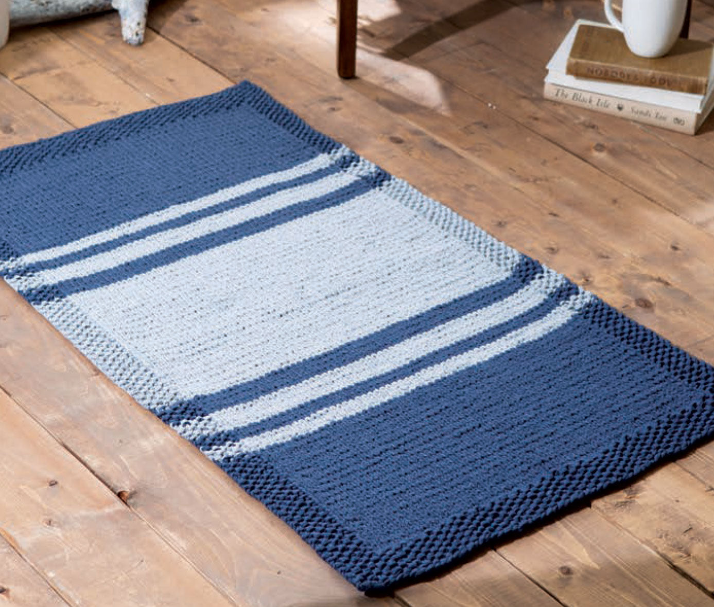 The Chunky Home Decor Digital Download by Leisure Arts features a knitted rectangular rug with a blue border and center, three light gray stripes near each end, perfect for adding cozy style to your space.