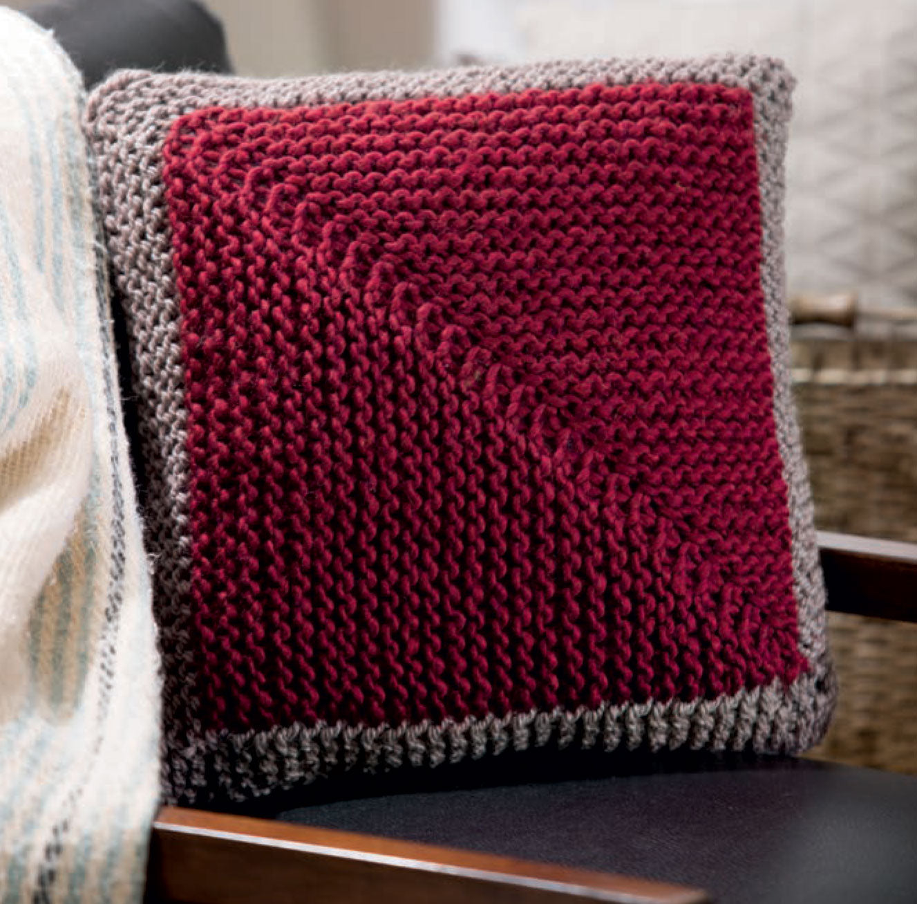 The Chunky Home Decor Digital Download by Leisure Arts features a knitted pillow with a deep red center, light gray border, and diagonal textured pattern—plus a nearby striped blanket—for a cozy touch to your space.