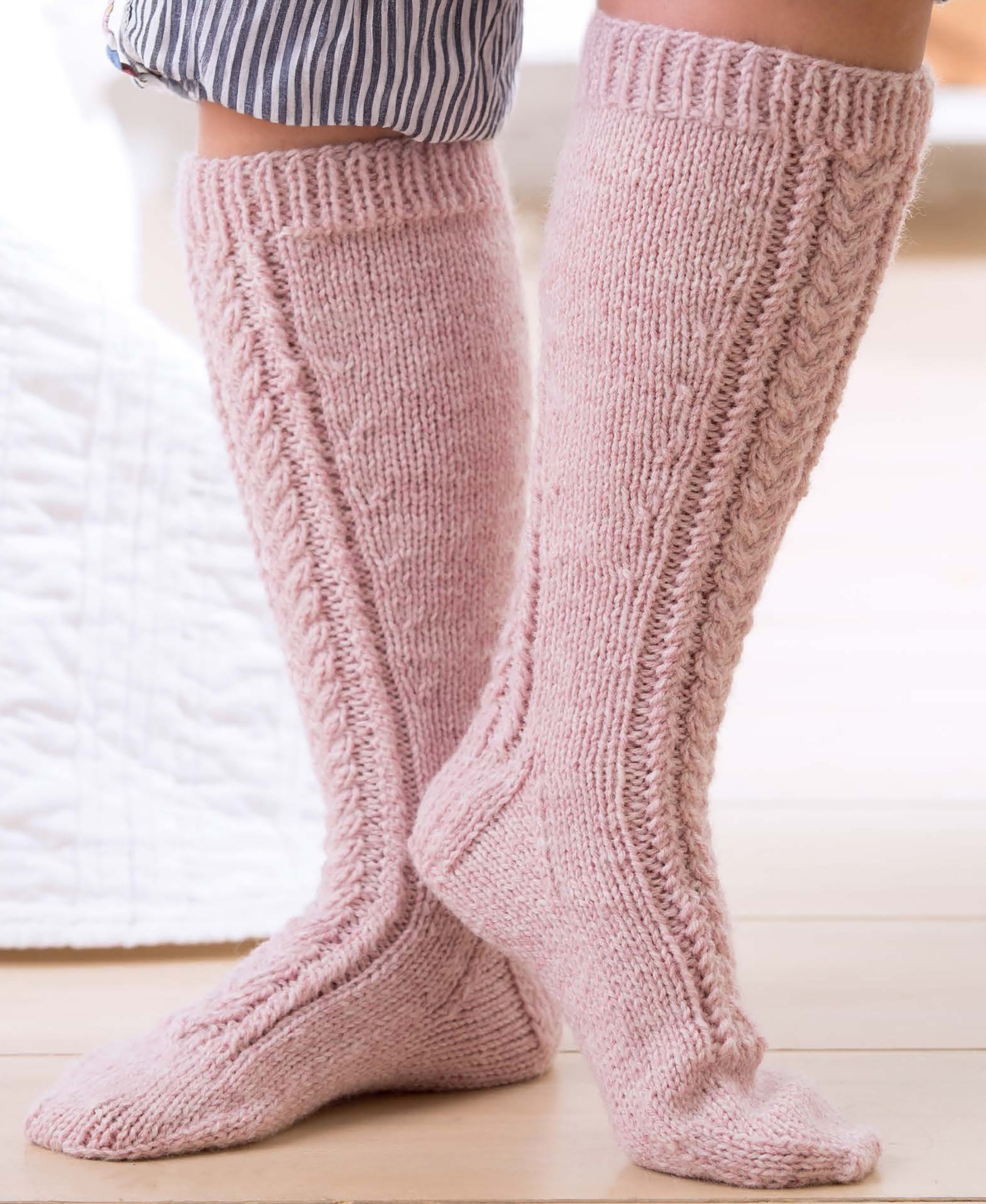 Wearing More Fun Socks by Leisure Arts, cozy knee-high light pink knit socks with a cable pattern, someone stands indoors on a light floor. These soft, warm socks are ideal for beginner knitting and pair perfectly with striped pajama pants.