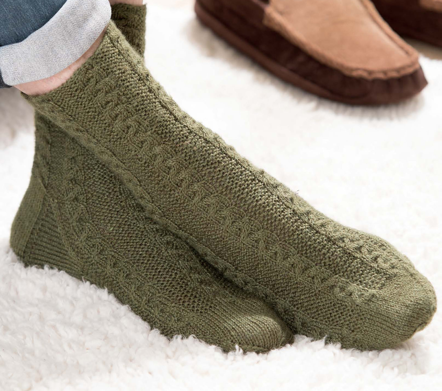 A person in green knit socks (More Fun Socks by Leisure Arts) sits on a fluffy white rug with rolled-up pants. Brown slippers appear in the background—great inspiration for cozy, beginner-friendly sock projects.