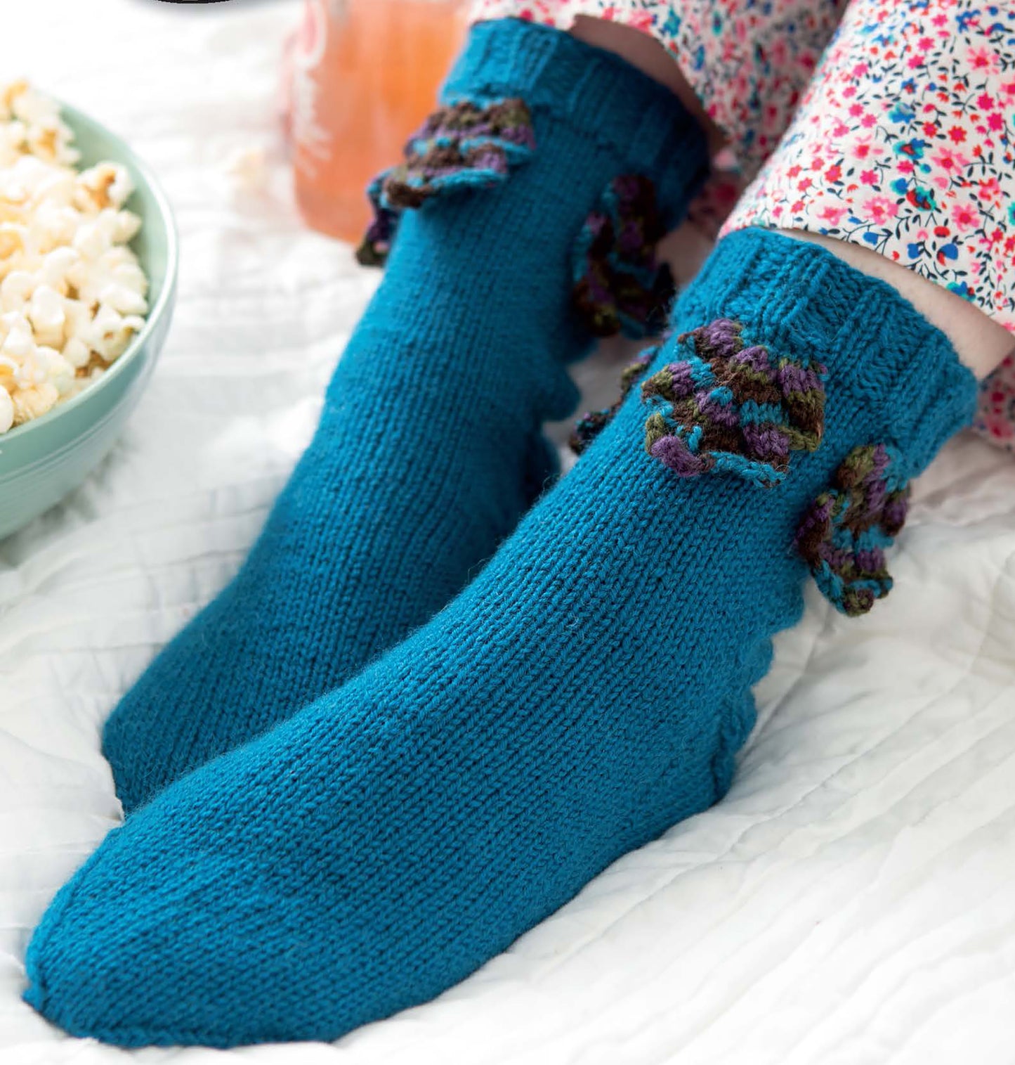 A person in floral pajama pants sits on a white blanket beside popcorn, wearing Leisure Arts’ More Fun Socks—bright blue knit socks with multicolored ruffles, ideal for beginner projects.