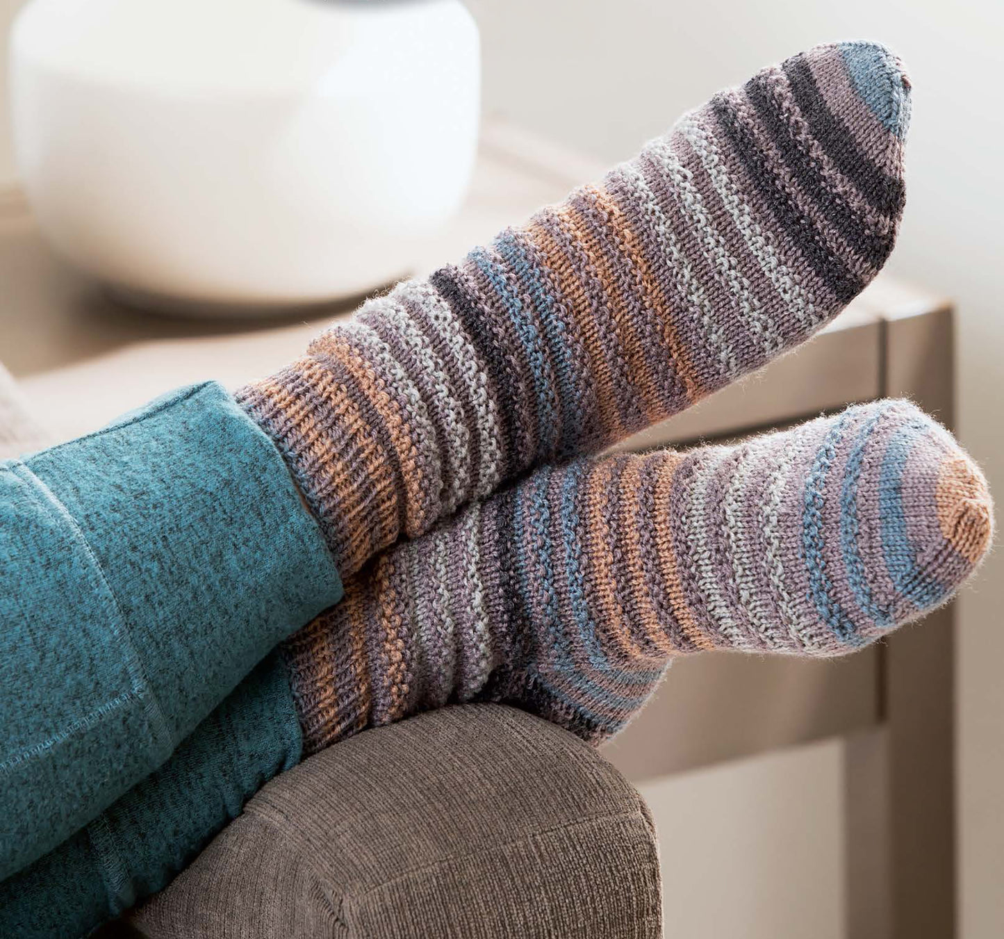A person in teal pants relaxes on a gray sofa, showing off Leisure Arts More Fun Socks—cozy, multicolored knit socks. A blurred white vase sits on a table in the background.