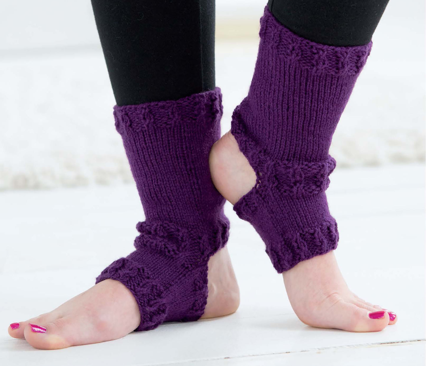 A person stands barefoot on a light floor, wearing black leggings and Leisure Arts’ More Fun Socks—purple knitted leg warmers perfect for beginners or trying out family sock patterns—with their heel and toes exposed.