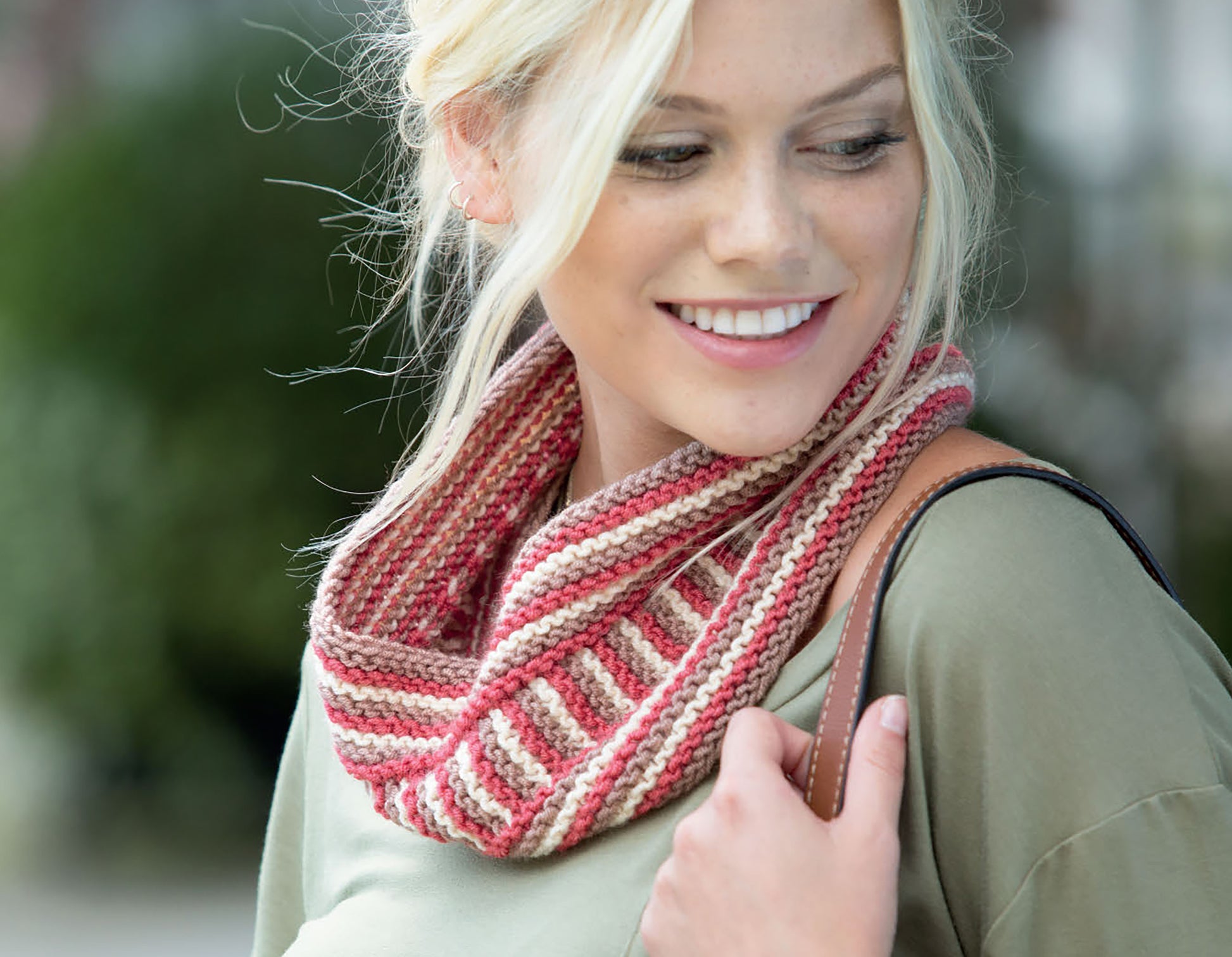 A blonde woman in a green top models a cozy, beginner knit cowl in red, pink, and beige from "Warm up with Scarves & Cowls" by Leisure Arts. She holds a brown shoulder bag and looks left, highlighting the stylish knitting design.