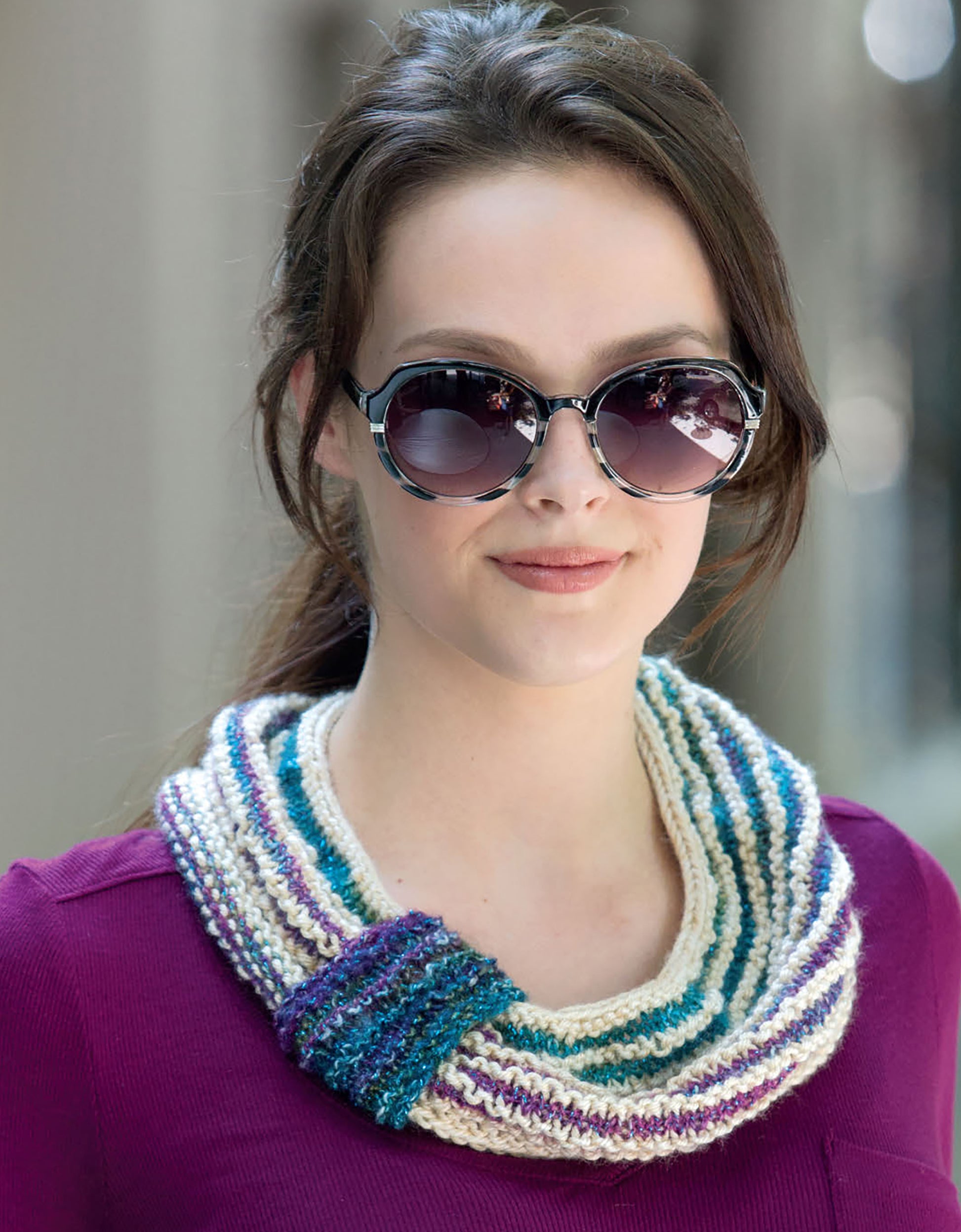 A woman in large round sunglasses, a purple top, and a multicolored cowl from "Warm up with Scarves & Cowls" by Leisure Arts, featuring cream, teal, and purple stripes, poses outdoors with a slight smile.