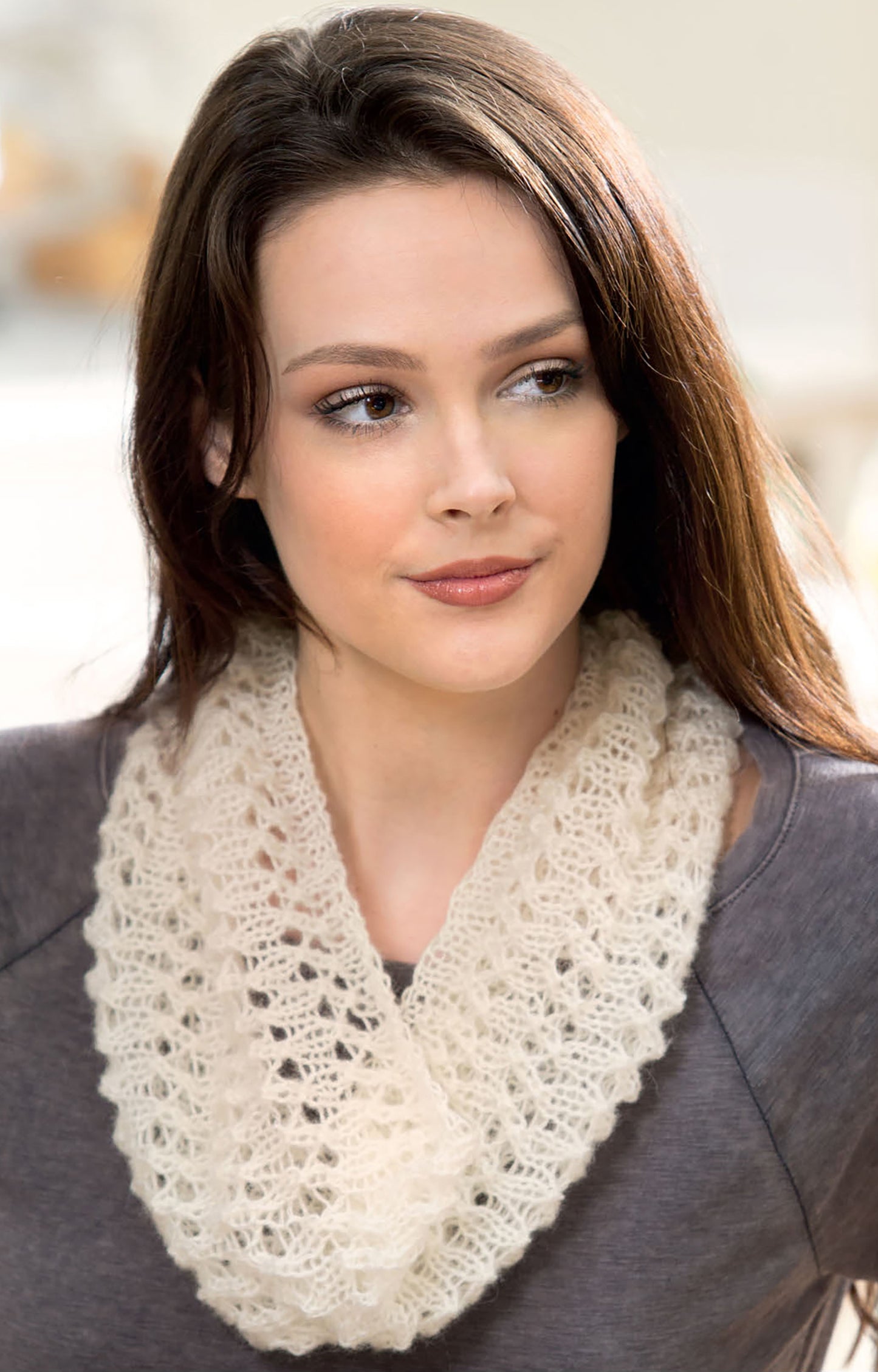 A woman with long brown hair in a grey top and cream lacy knit scarf from "Warm up with Scarves & Cowls" by Leisure Arts gazes calmly to the side, showcasing this elegant piece for those seeking new knitting designs.