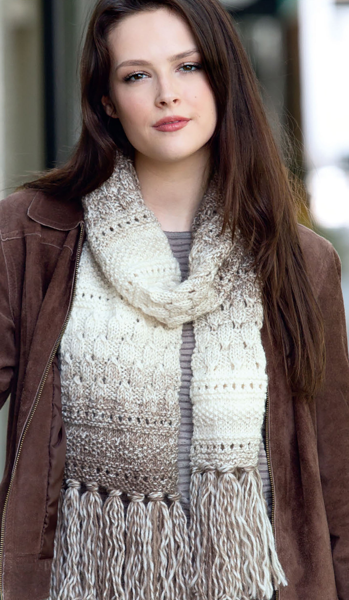 A woman with long brown hair styles a Leisure Arts "Warm up with Scarves & Cowls" chunky knit scarf in cream and brown with textured patterns and fringe, layered over a striped shirt and brown jacket.