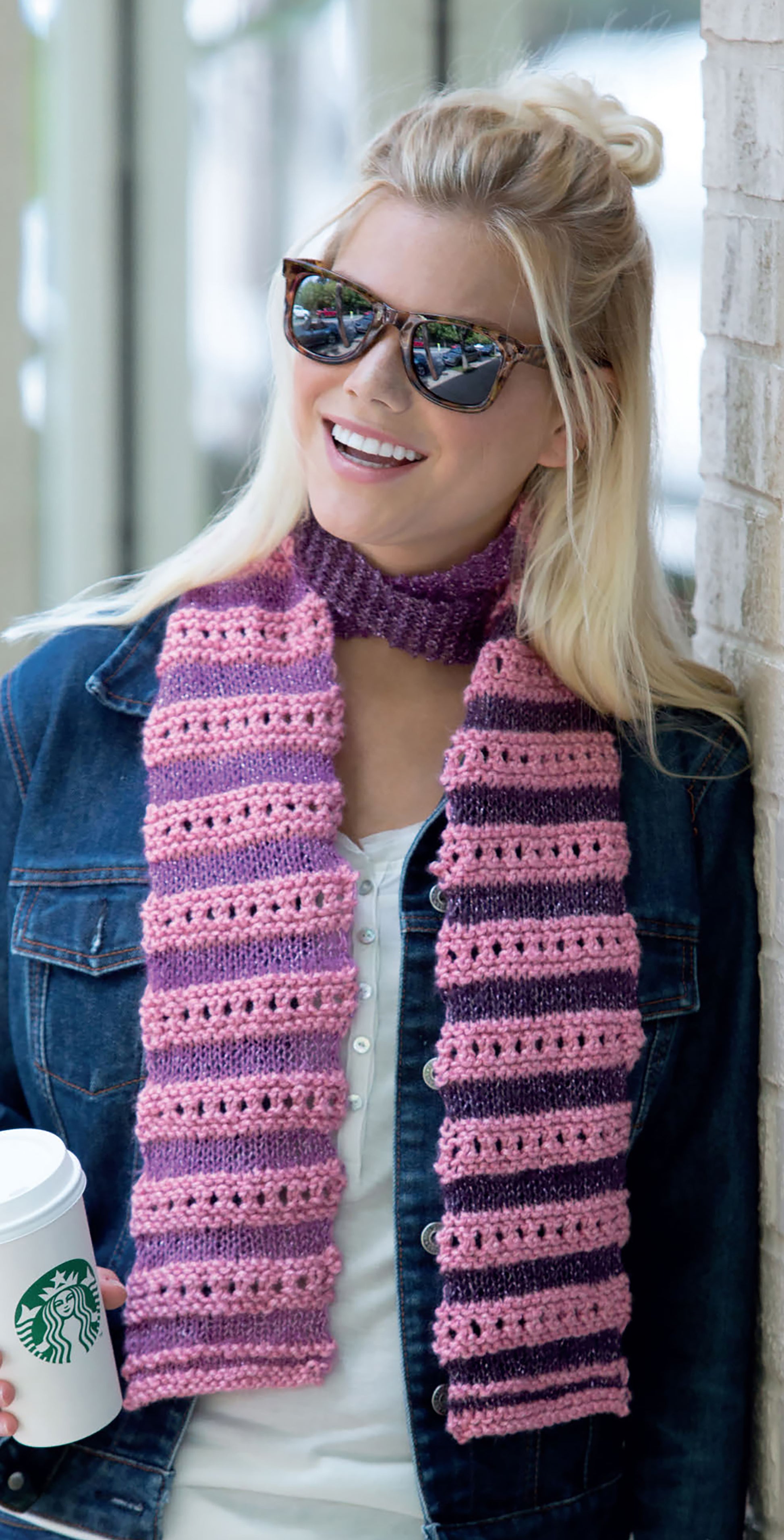 A woman in sunglasses, a denim jacket, and a pink striped scarf from "Warm up with Scarves & Cowls" by Leisure Arts leans against a wall, smiling with a Starbucks cup in hand.