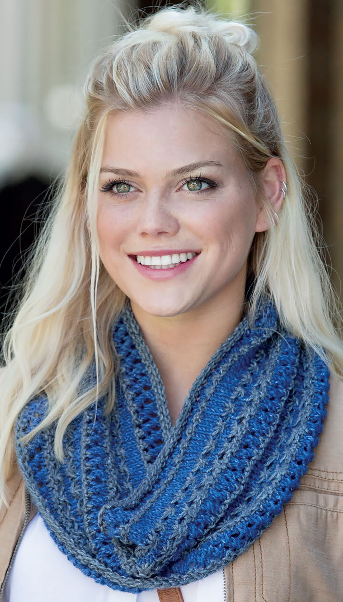 A smiling woman wears a blue and gray crocheted infinity scarf from "Warm up with Scarves & Cowls" by Leisure Arts, paired with a light brown jacket, standing outdoors against a softly blurred background.